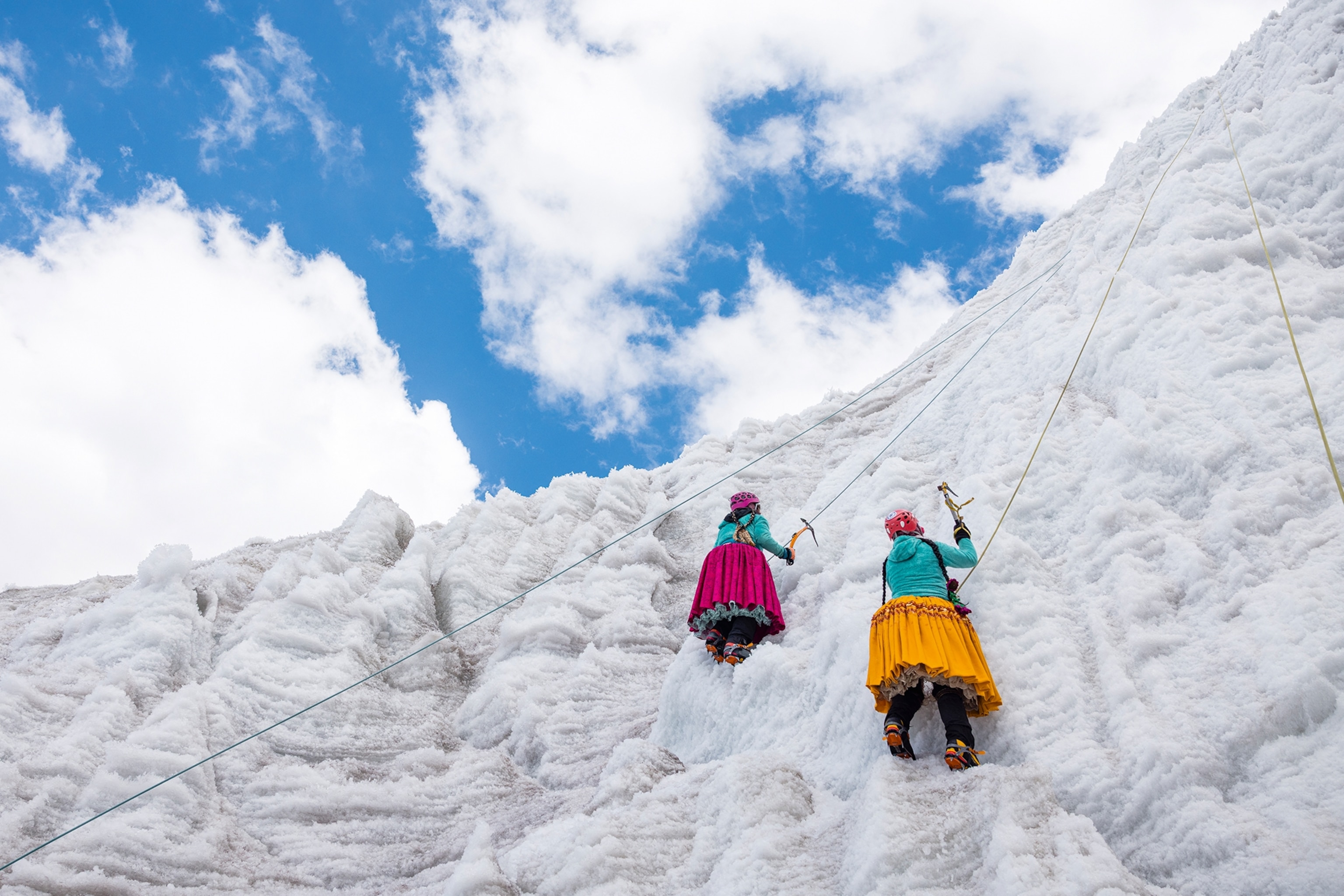 Two Indigenous women in puffy skirts climbing an ice wall in the mountains.