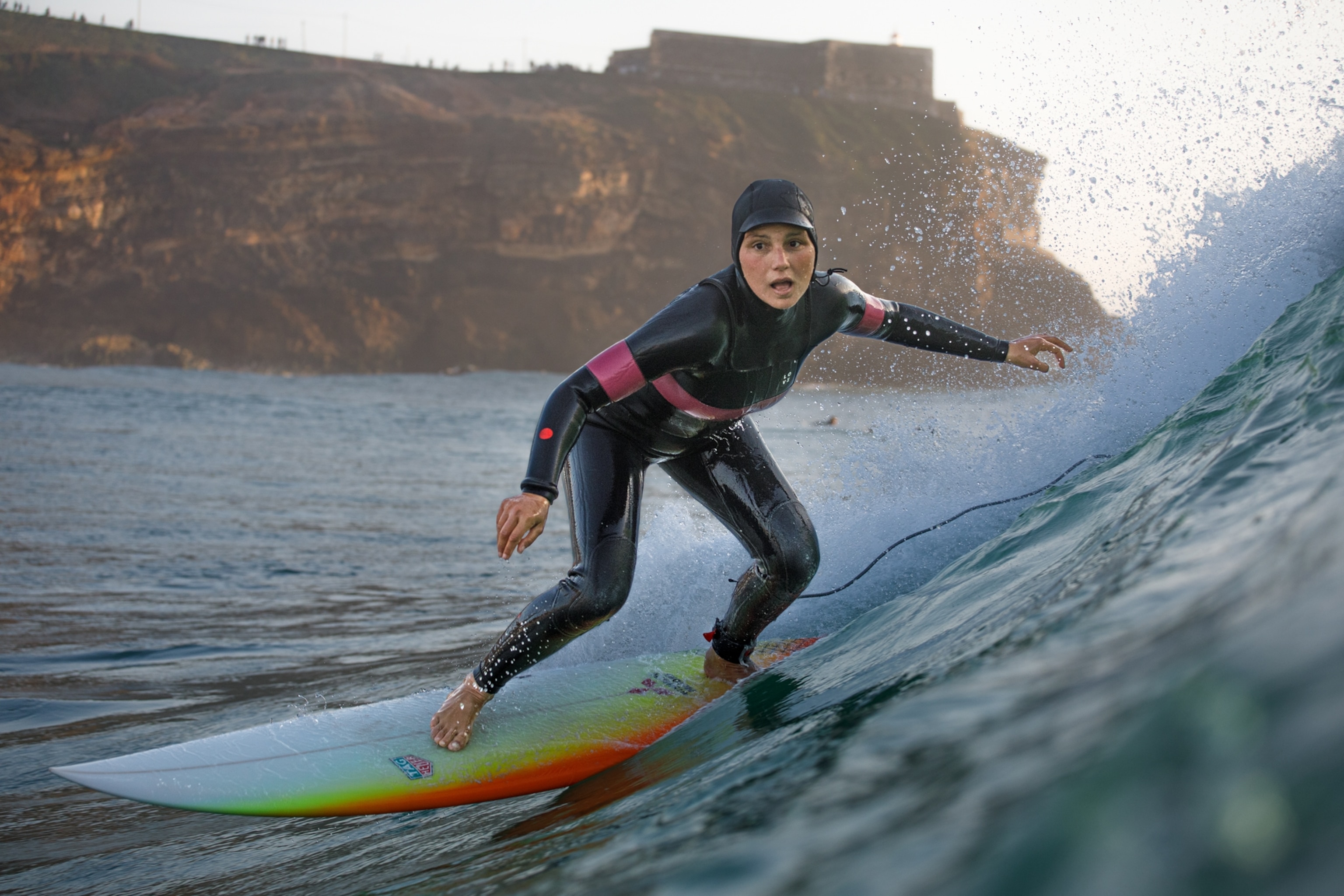 A woman in a wet suit is seen surfing on a wave. She is facing the camera and appears focused. There are rocky cliffs in the background.