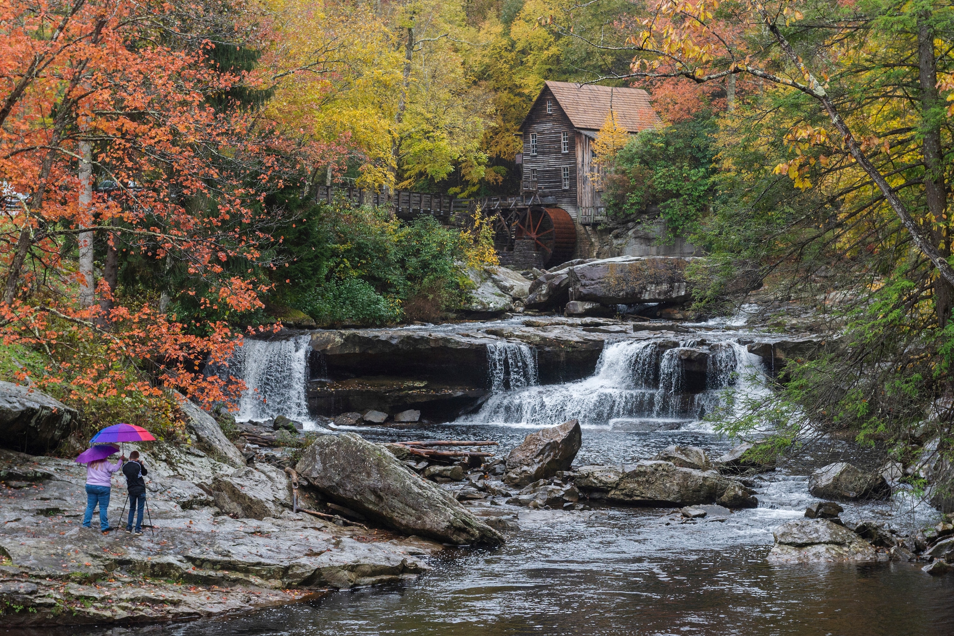 Two people stand under an umbrella and look at the waterfall in front of them, surrounded by colorful fall leaves and a cabin tucked away in the trees