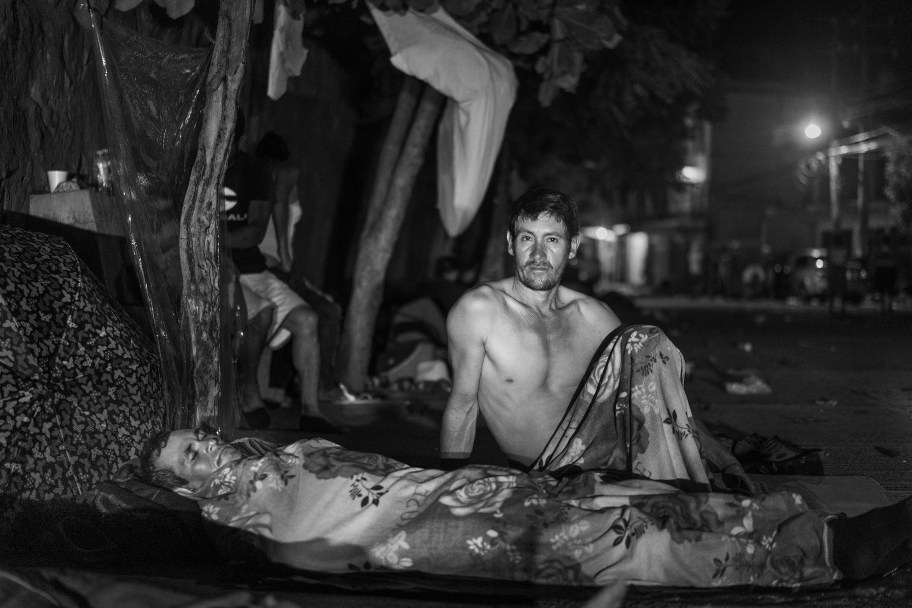 a shirtless man sitting and a man laying down covered by a blanket at night outside