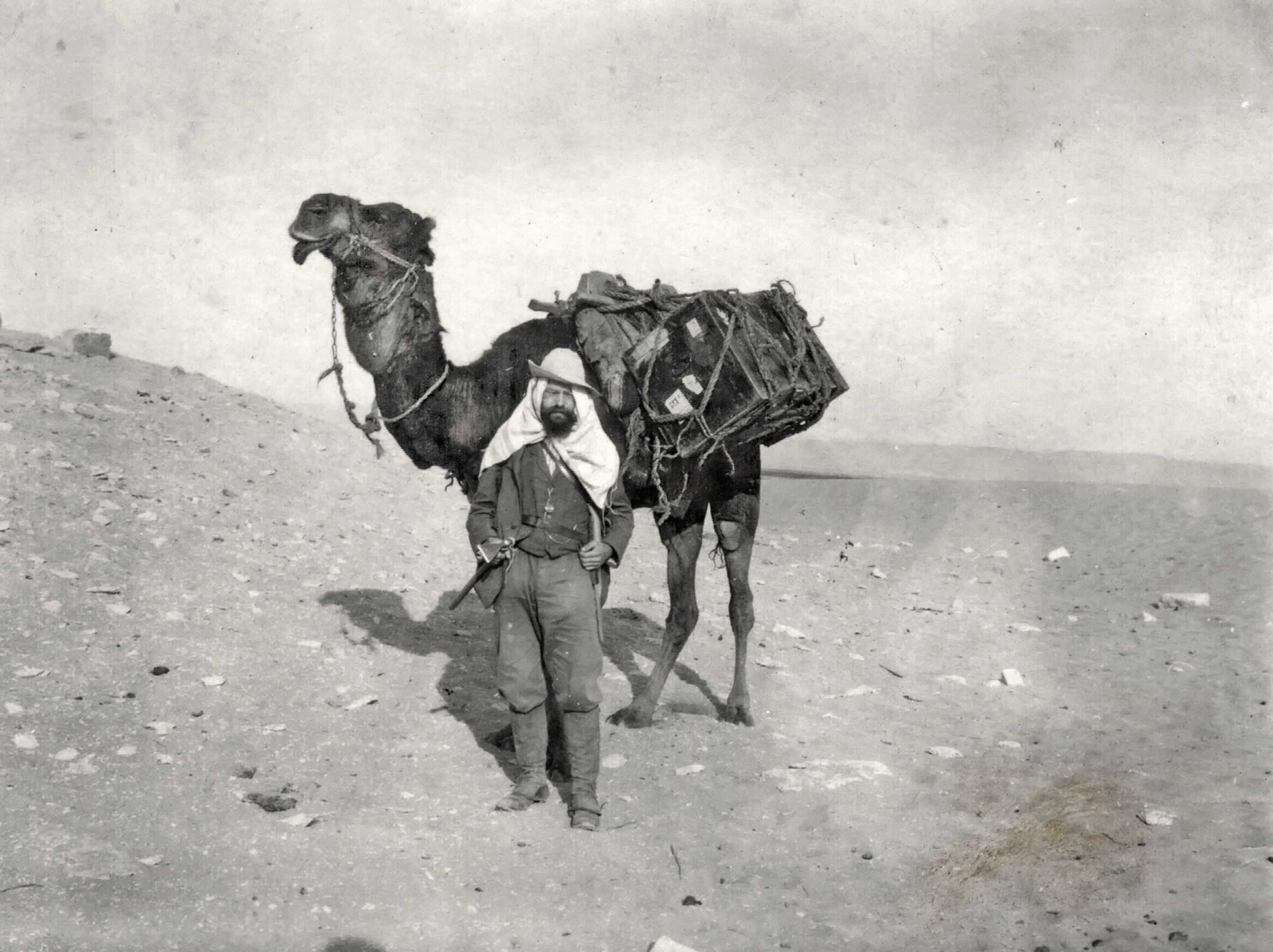 A bearded man in a hat and headscarf stands in a desert next to a camel carrying large, strapped-down wooden crates on its back in this vintage photo.