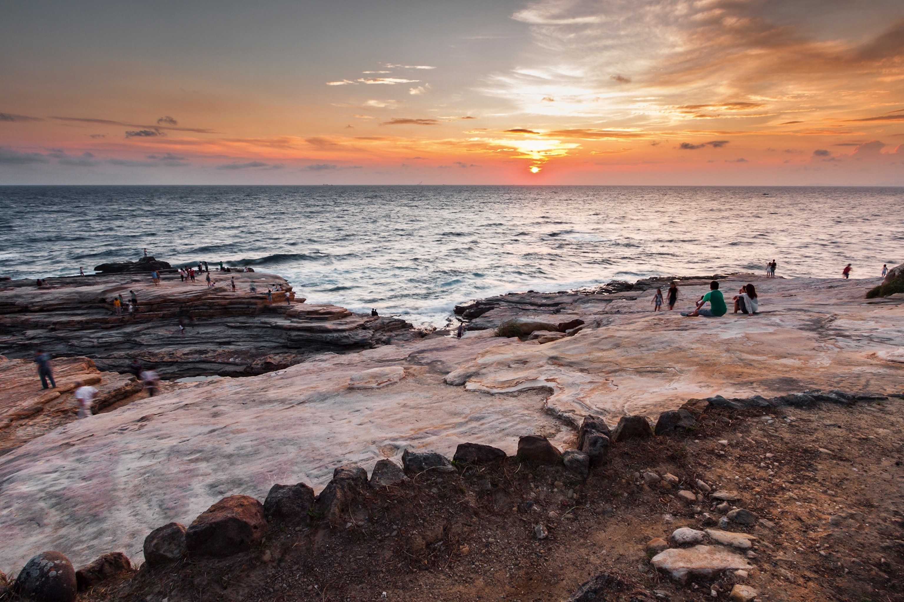 A wide, scenic shot of a rocky beach front with locals watching the sunset over the horizon.