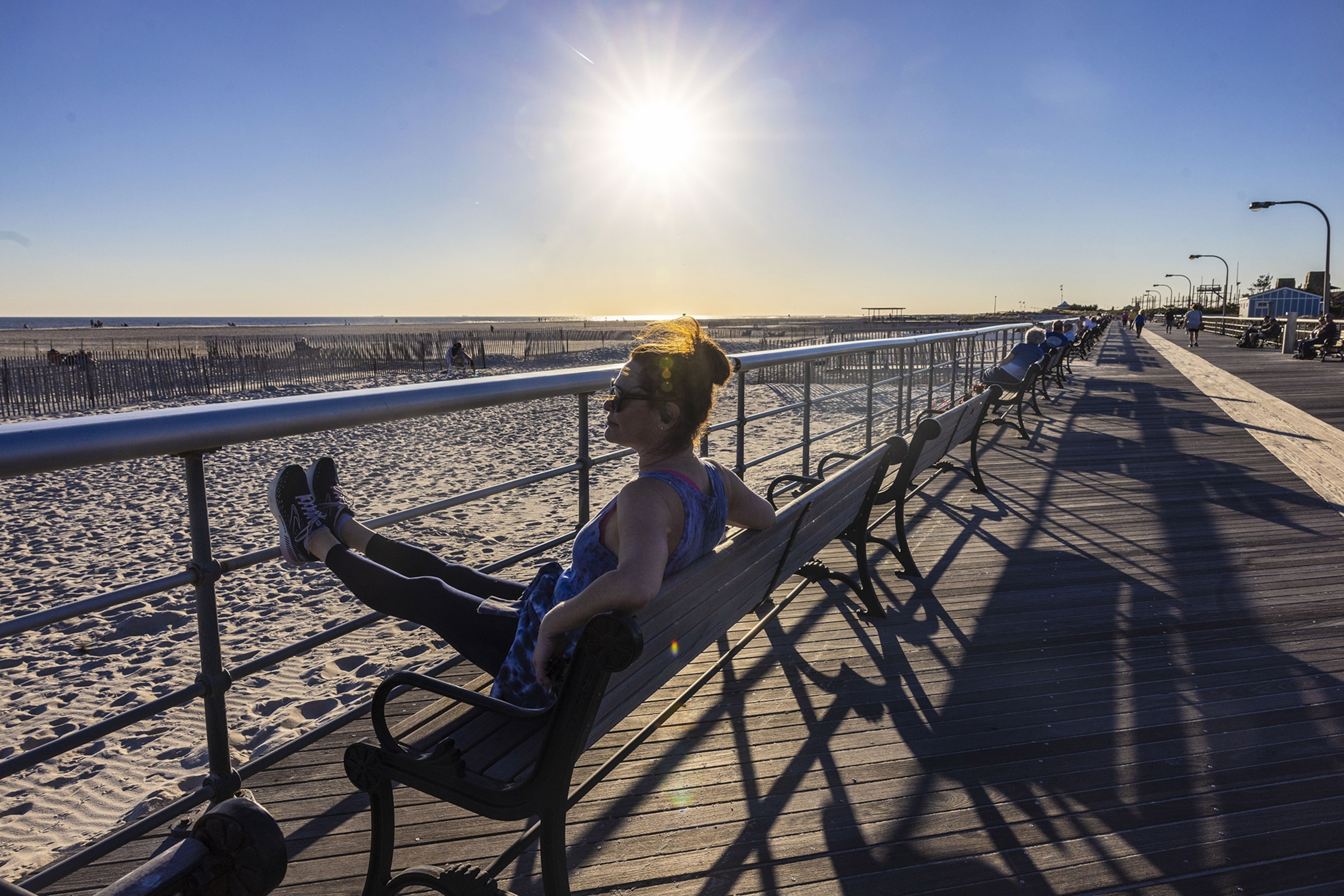 A woman sits on a bench on a boardwalk with her feet resting on railings, looking out at the beach and ocean