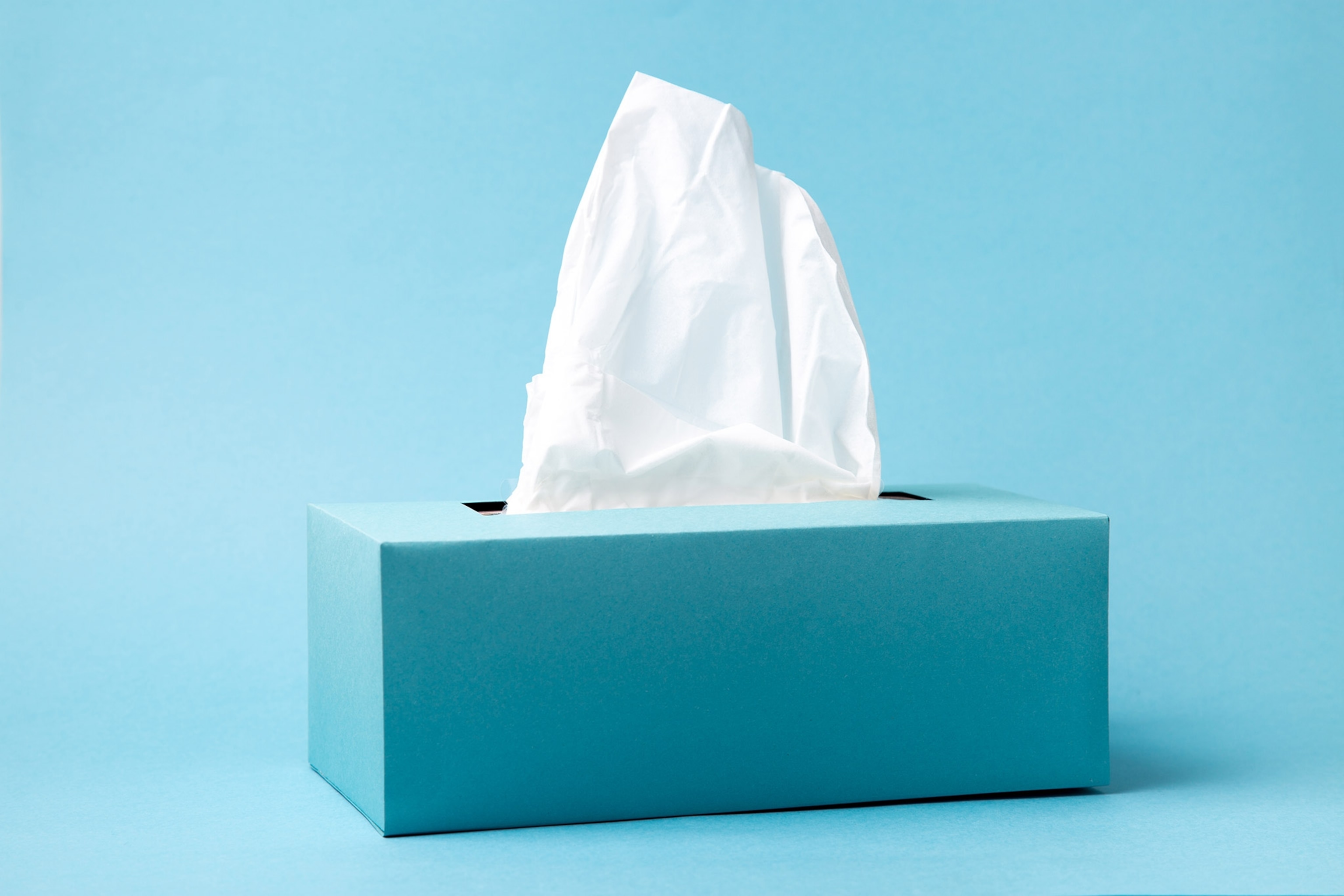 Blue facial tissue box with white tissue coming out the top, against blue background