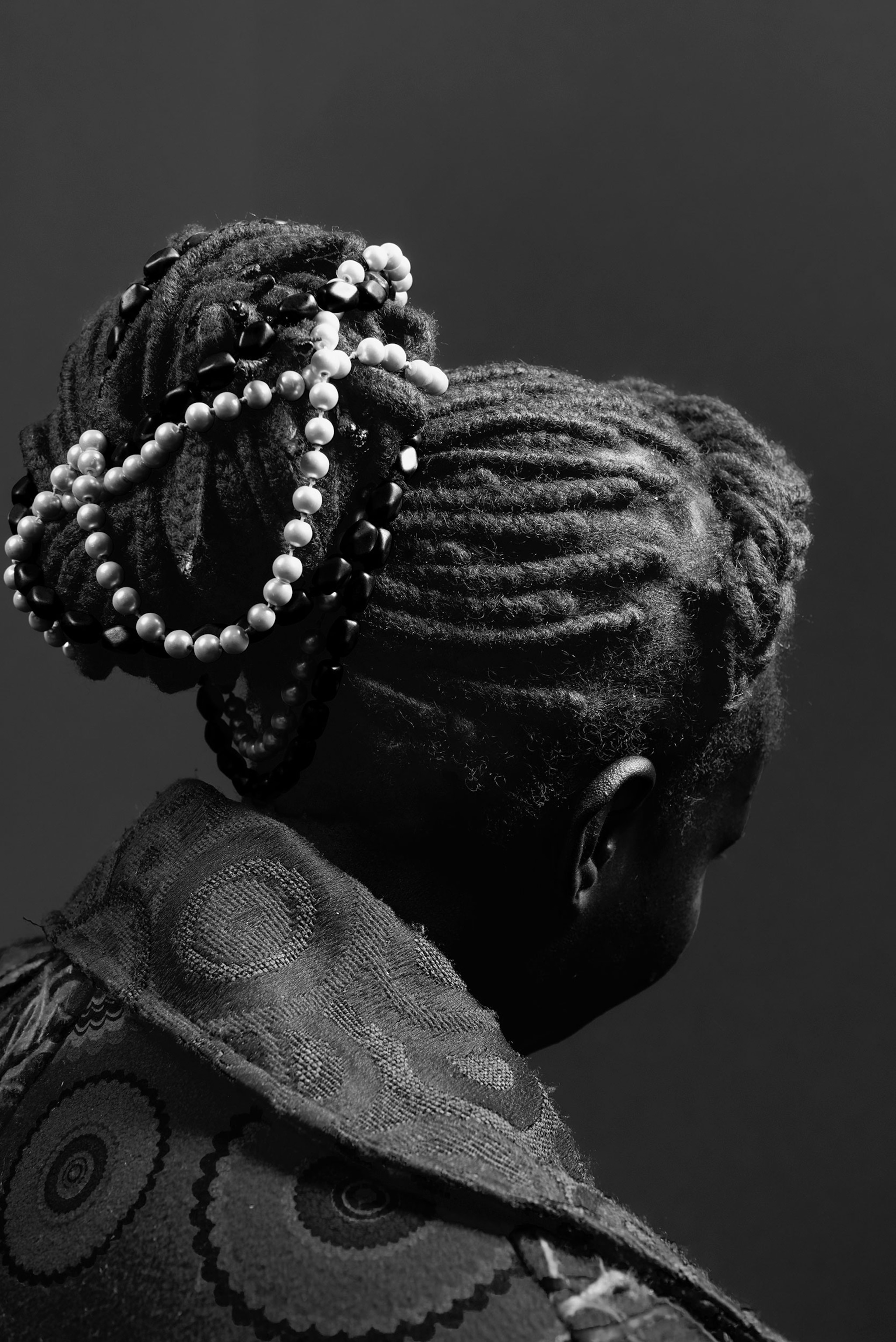 the back of a woman's head, her hair elaborately braided into a bun and adorned with strings of beads
