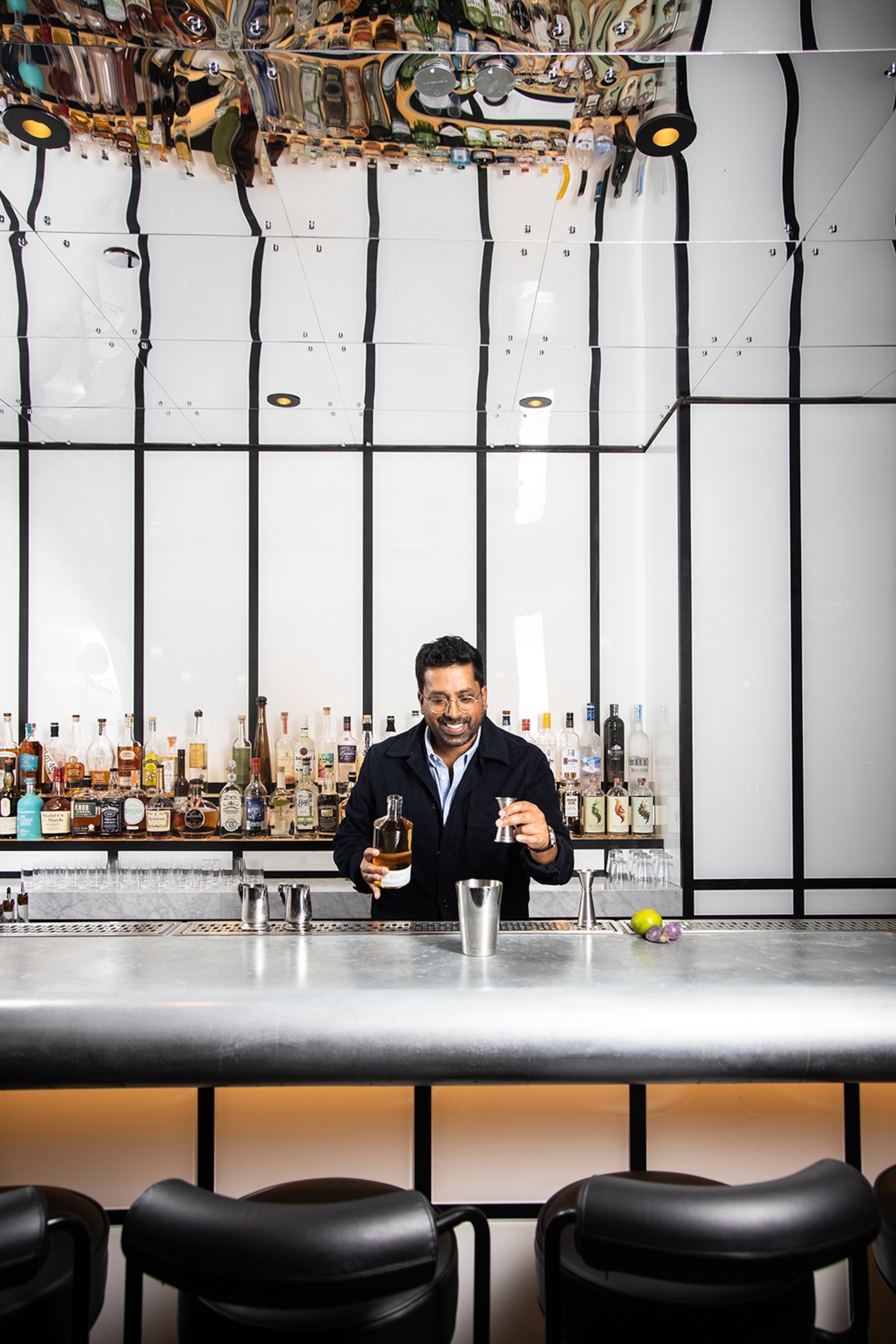 A dapper bartender of Indian heritage behind a sleek, futuristic bar with a mirrored ceiling.