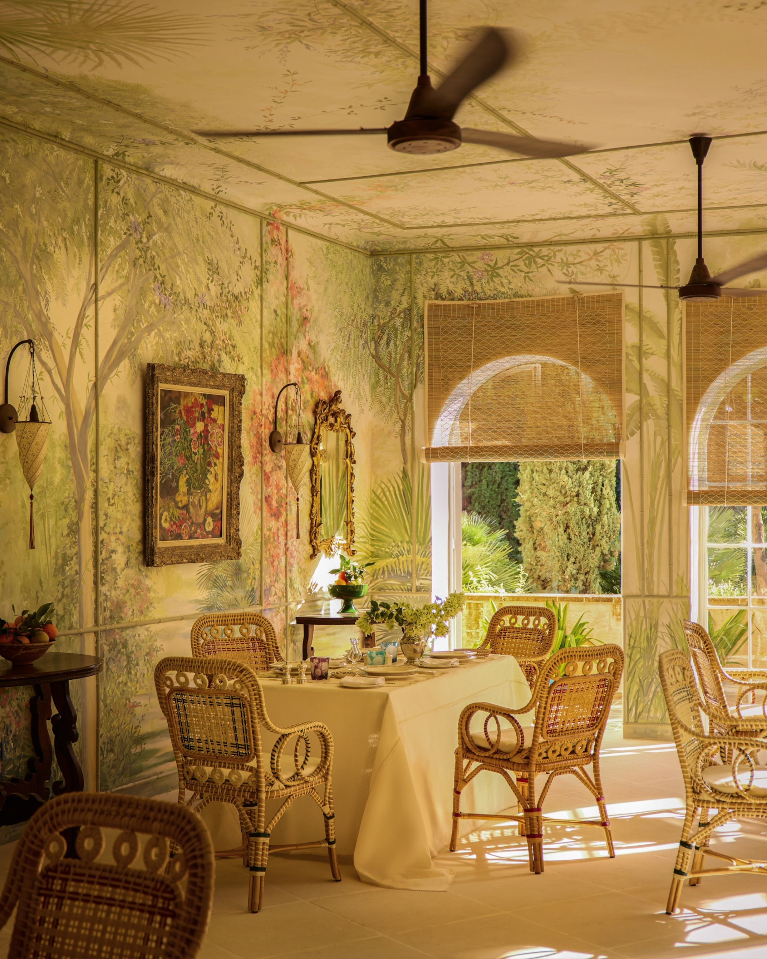 An elegant dining room with wicker chairs, botanical wall murals, ceiling fans, and large windows letting in warm sunlight.