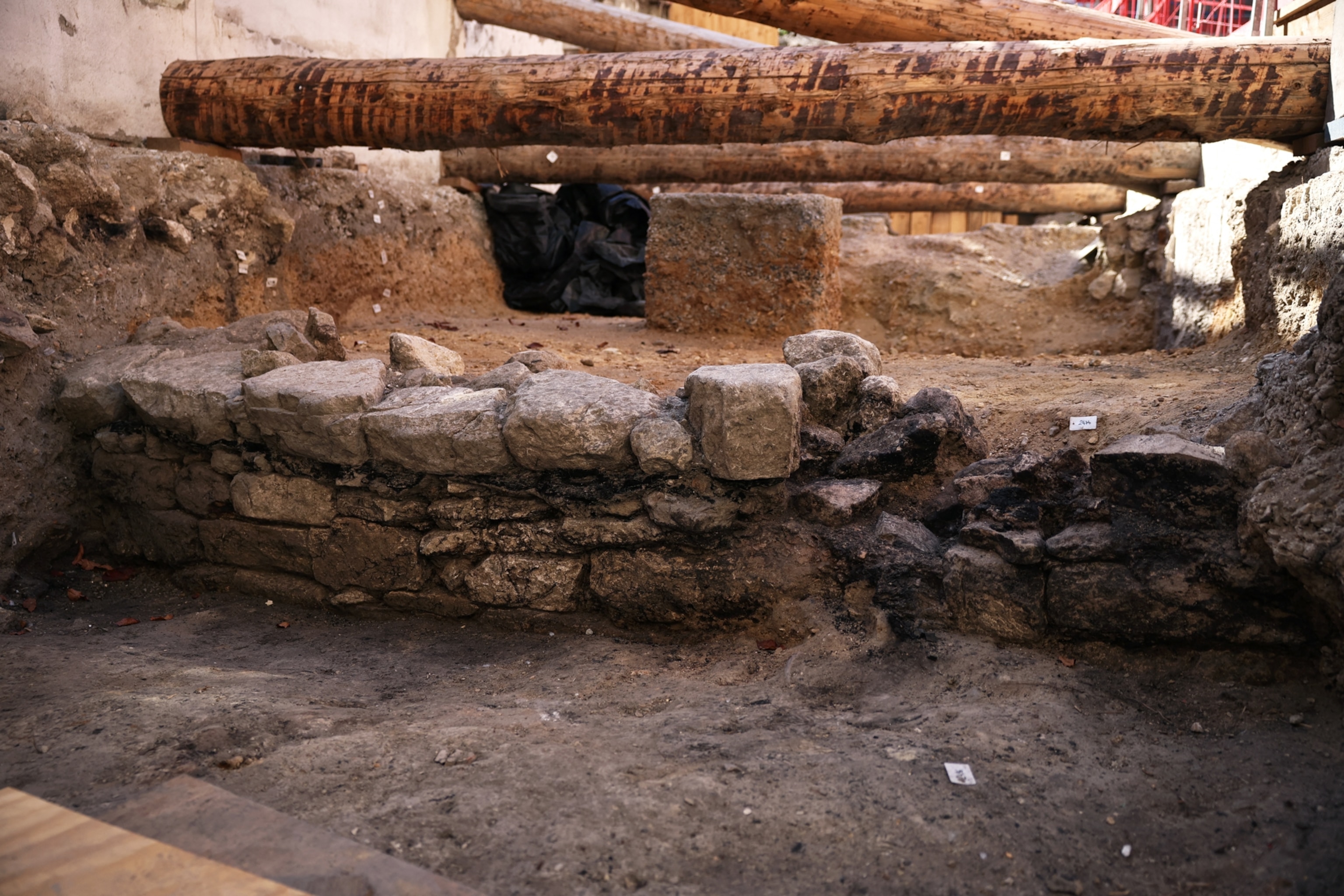 An archaeological dig reveals the remains of a low stone wall and building foundation, with wooden support beams spanning the excavation trench above.