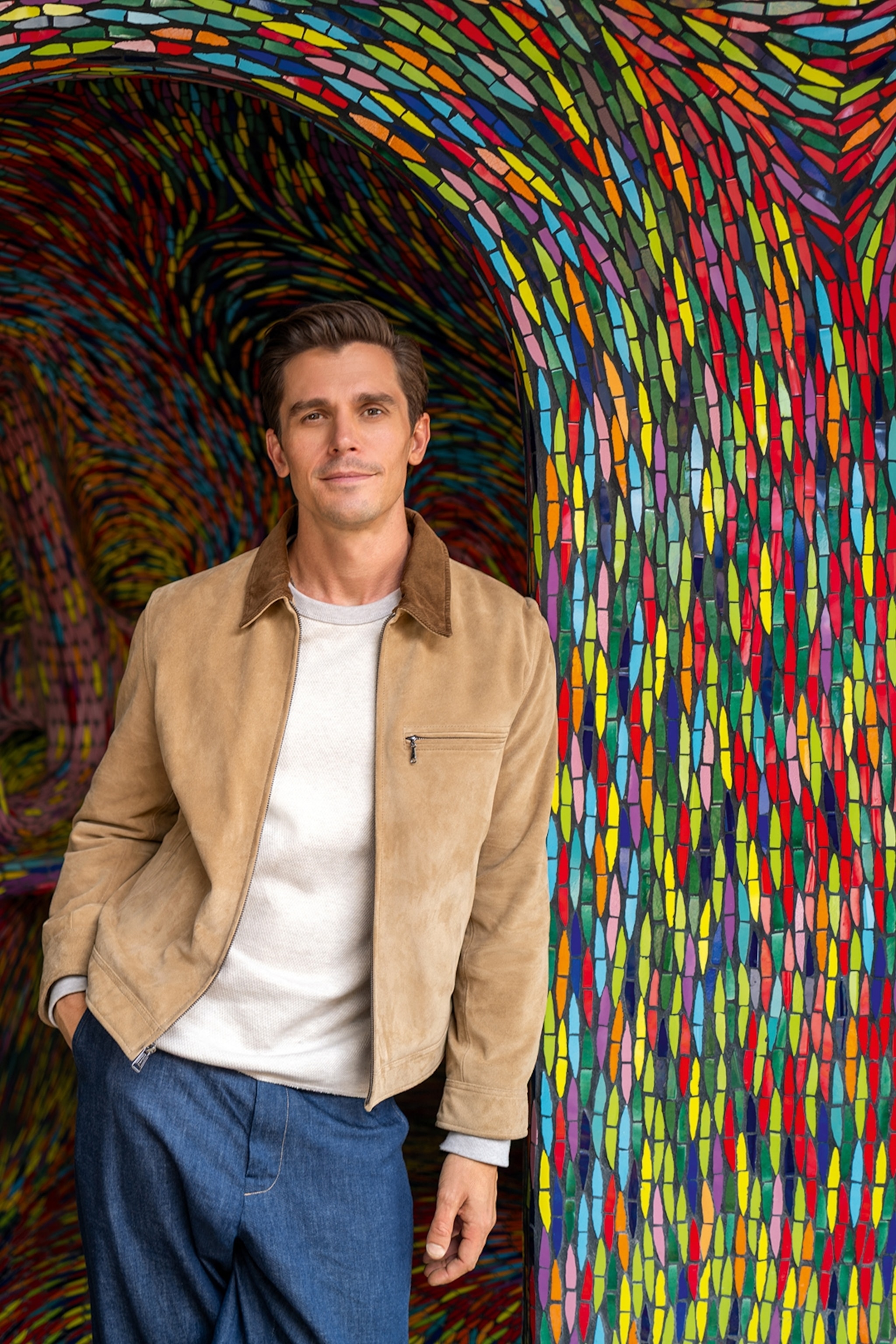 Portrait of a young man with stylish clothes casually leaning against a curved mosaic wall.