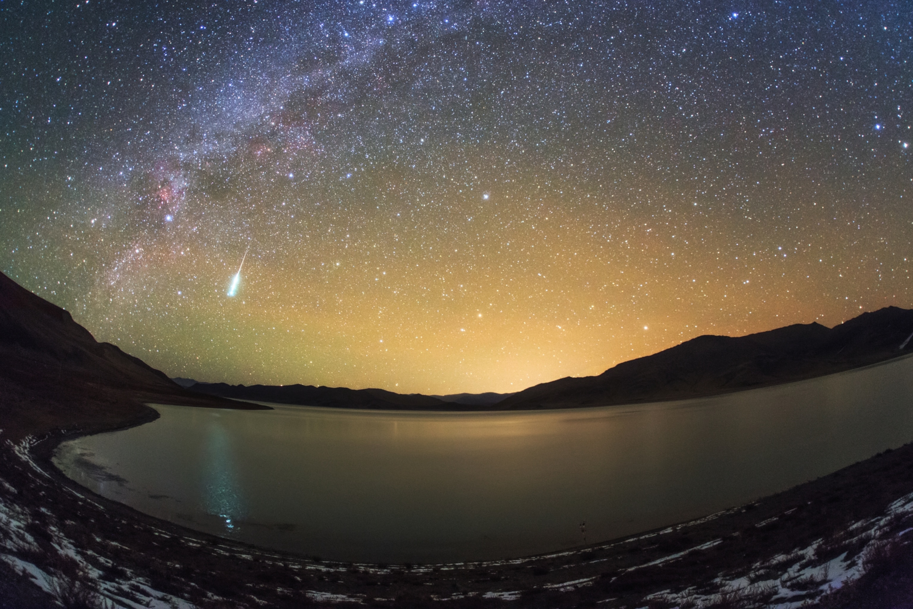 The white streak of a meteor in a star-filled sky, above a lake and low snowy mountains