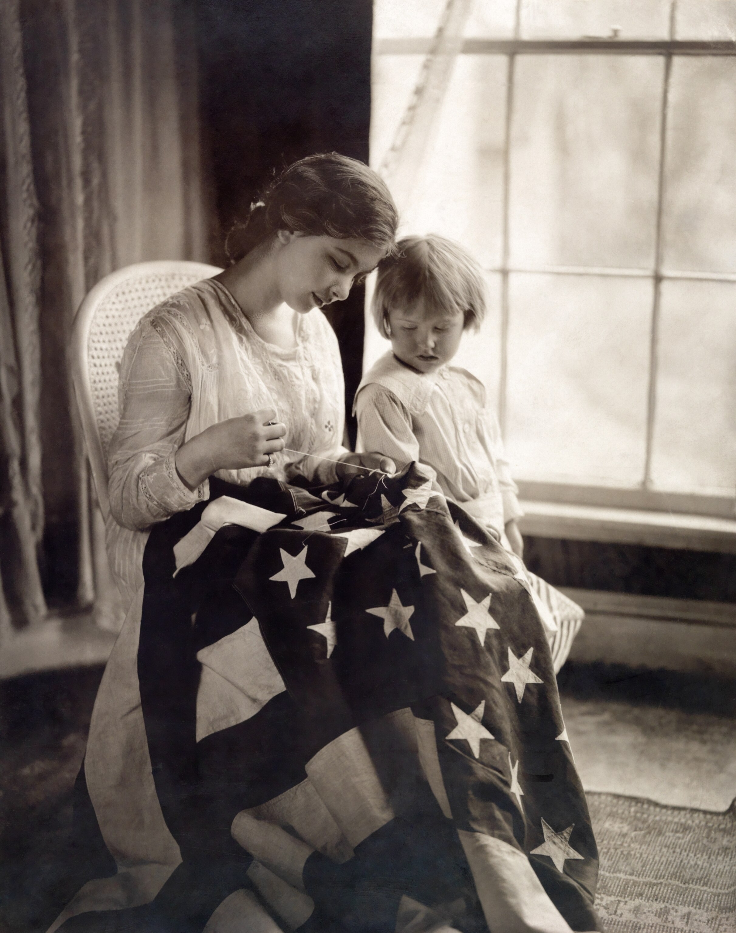 A woman sews an American flag as a young girl looks on, 1917.