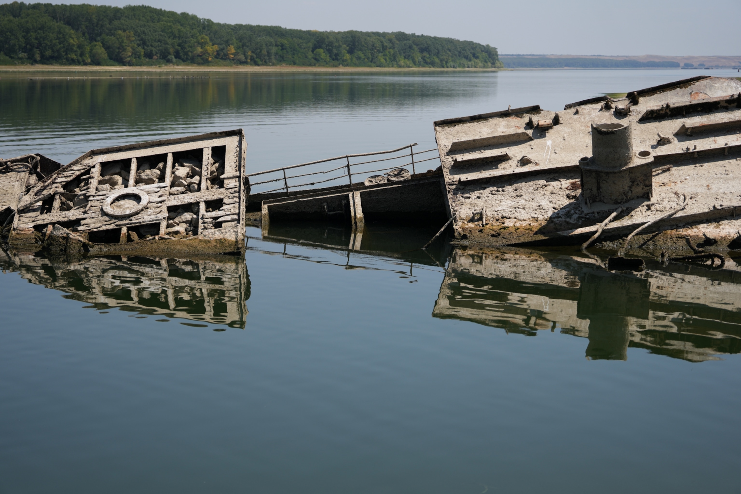 The rusted, broken remains of a sunken metal ship emerge from calm, reflective river water, with a dense green forest lining the distant shore.
