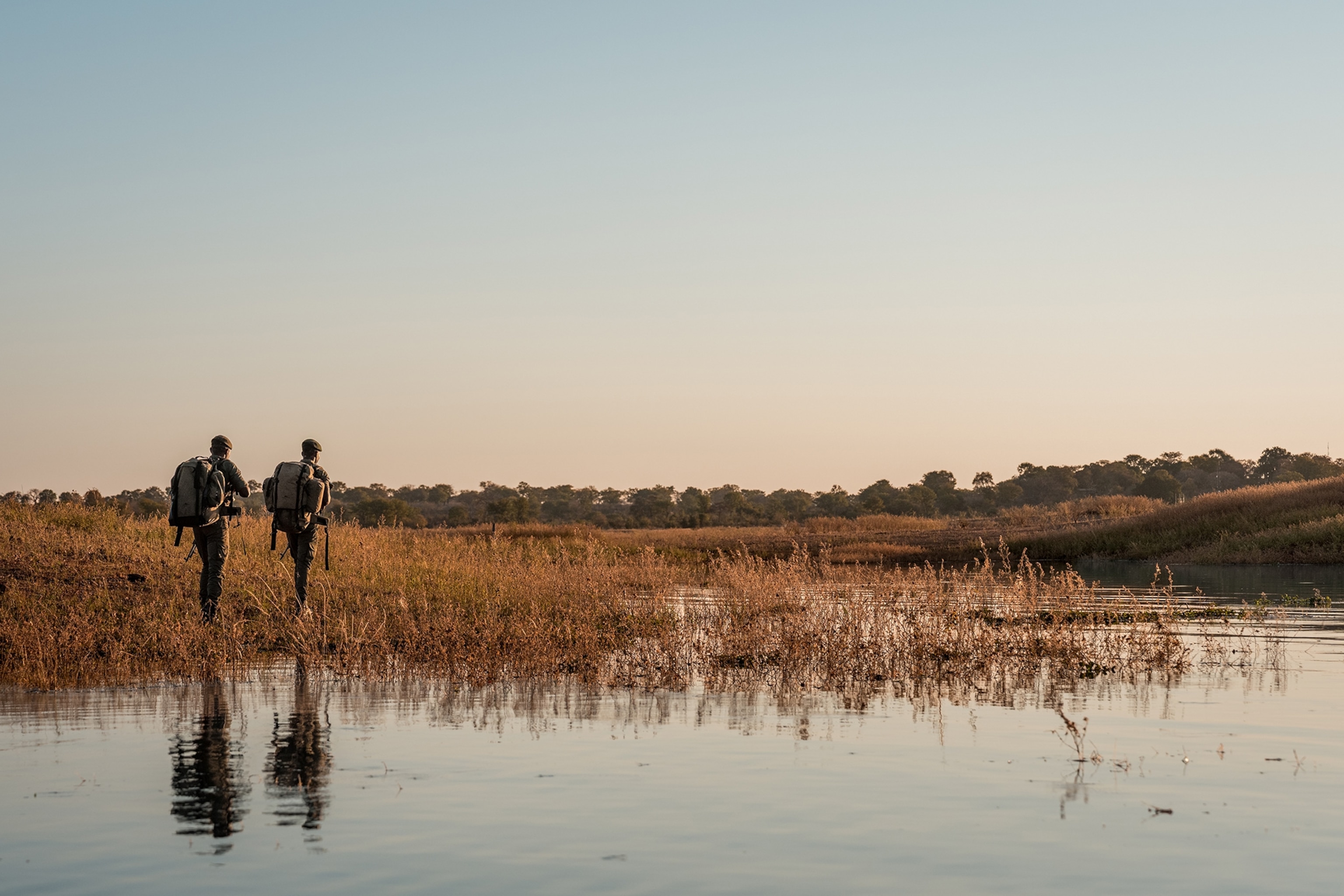 two park rangers on patrol seen from behind walking on the shore of a lake. Their reflections are seen in the water.
