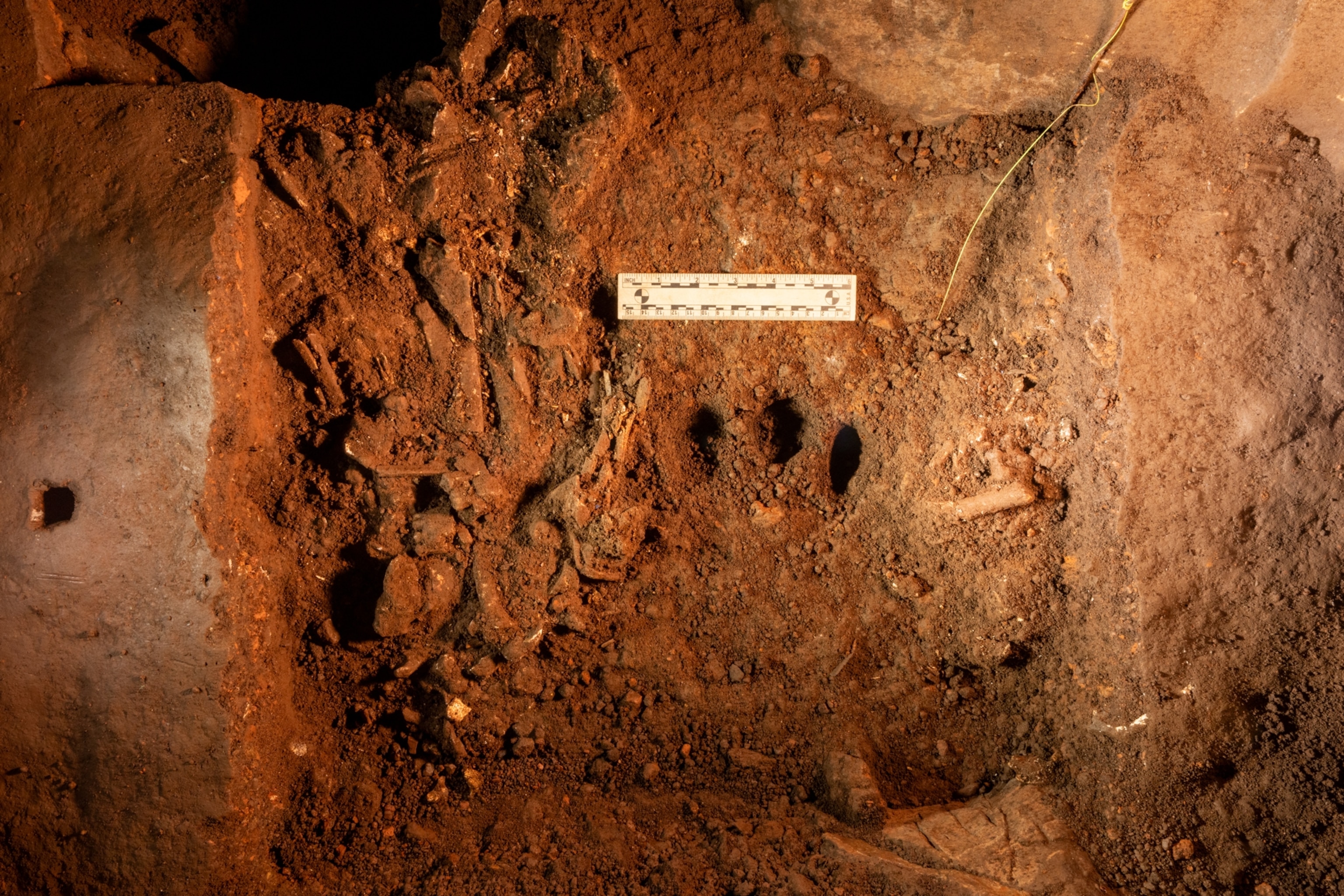an archaeological dig site with red-brown soil and a three small holes. A scale ruler is placed for measurement.