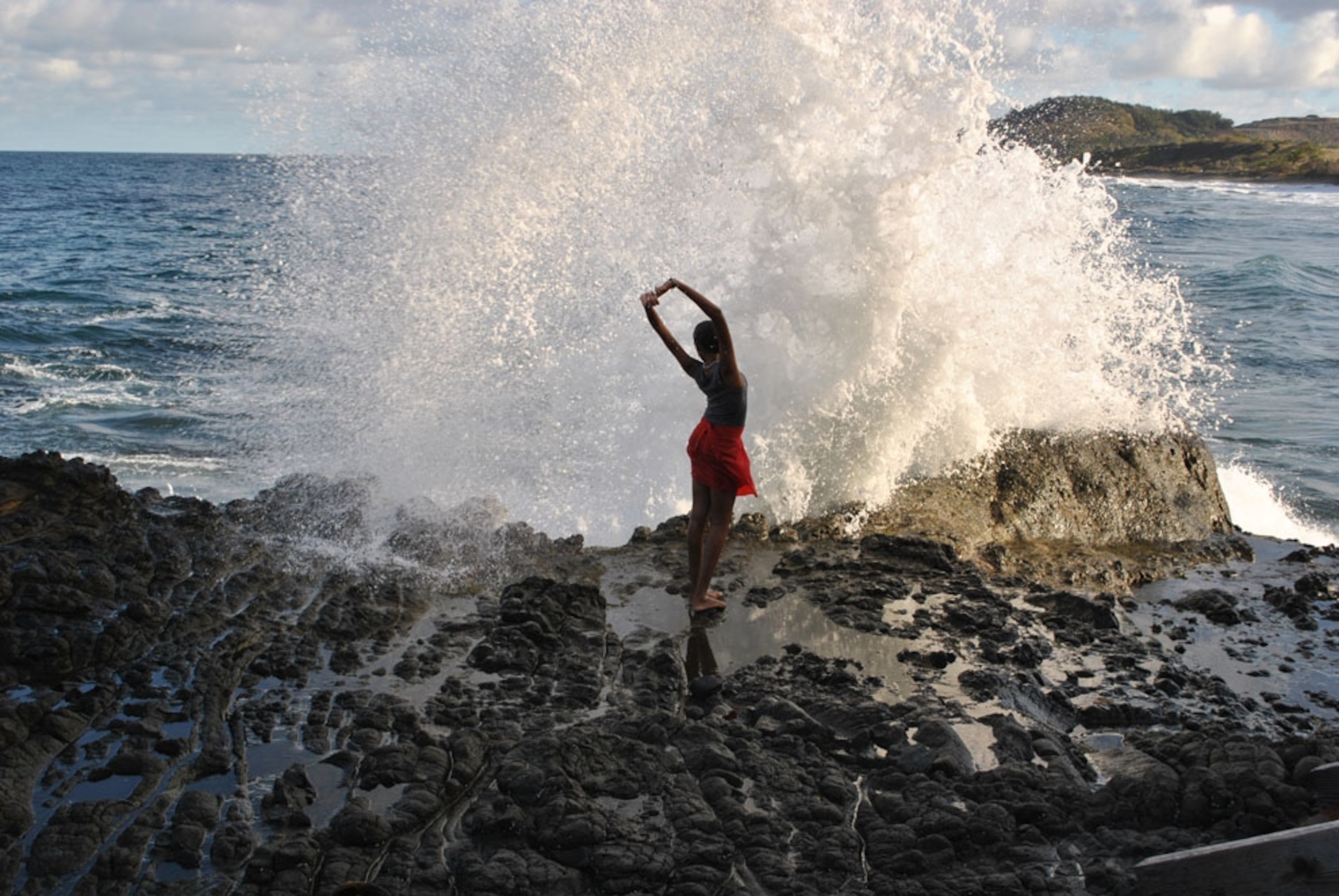 A woman poses as a wave crashes against a rocky ledge in the Caribbean Isles