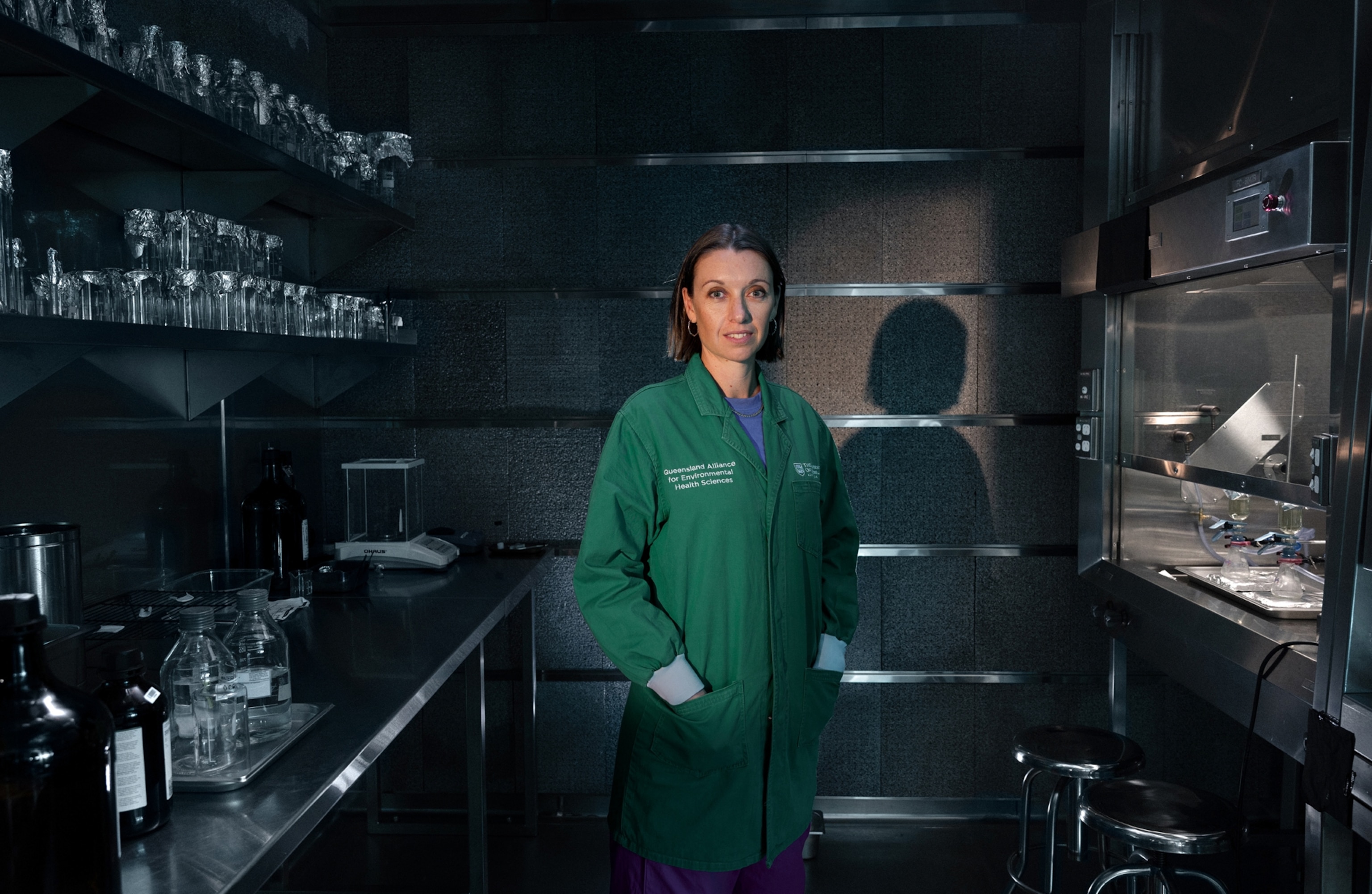 A woman stands for a portrait in a small dark room with beakers and other equipment around her.