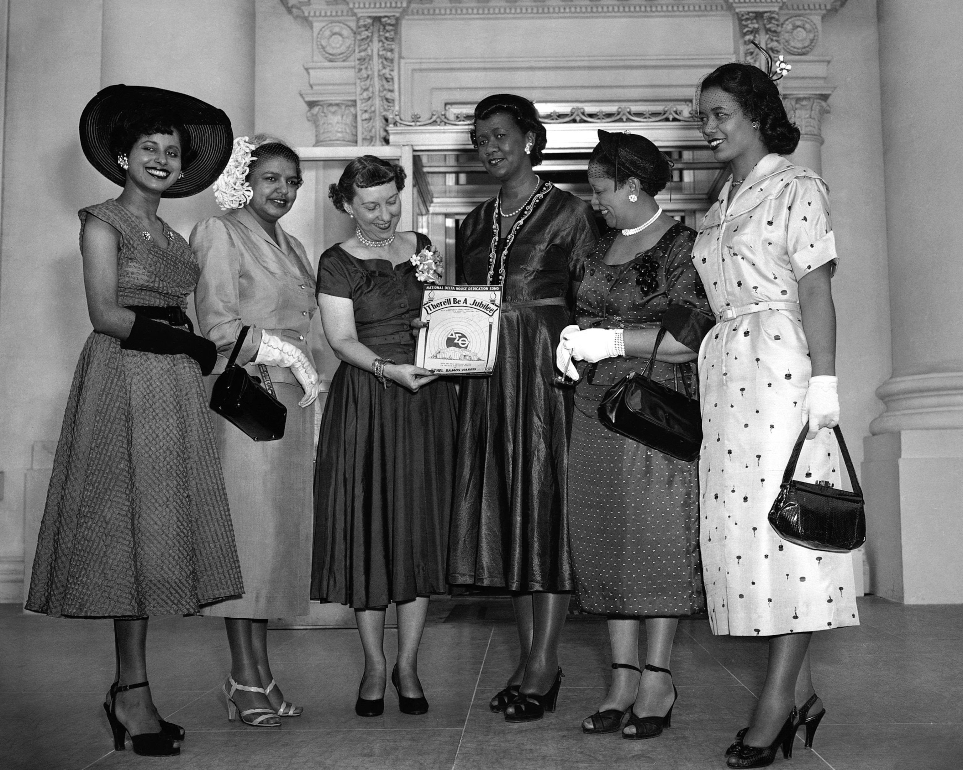 Six elegantly dressed women, holding a certificate together, smiling