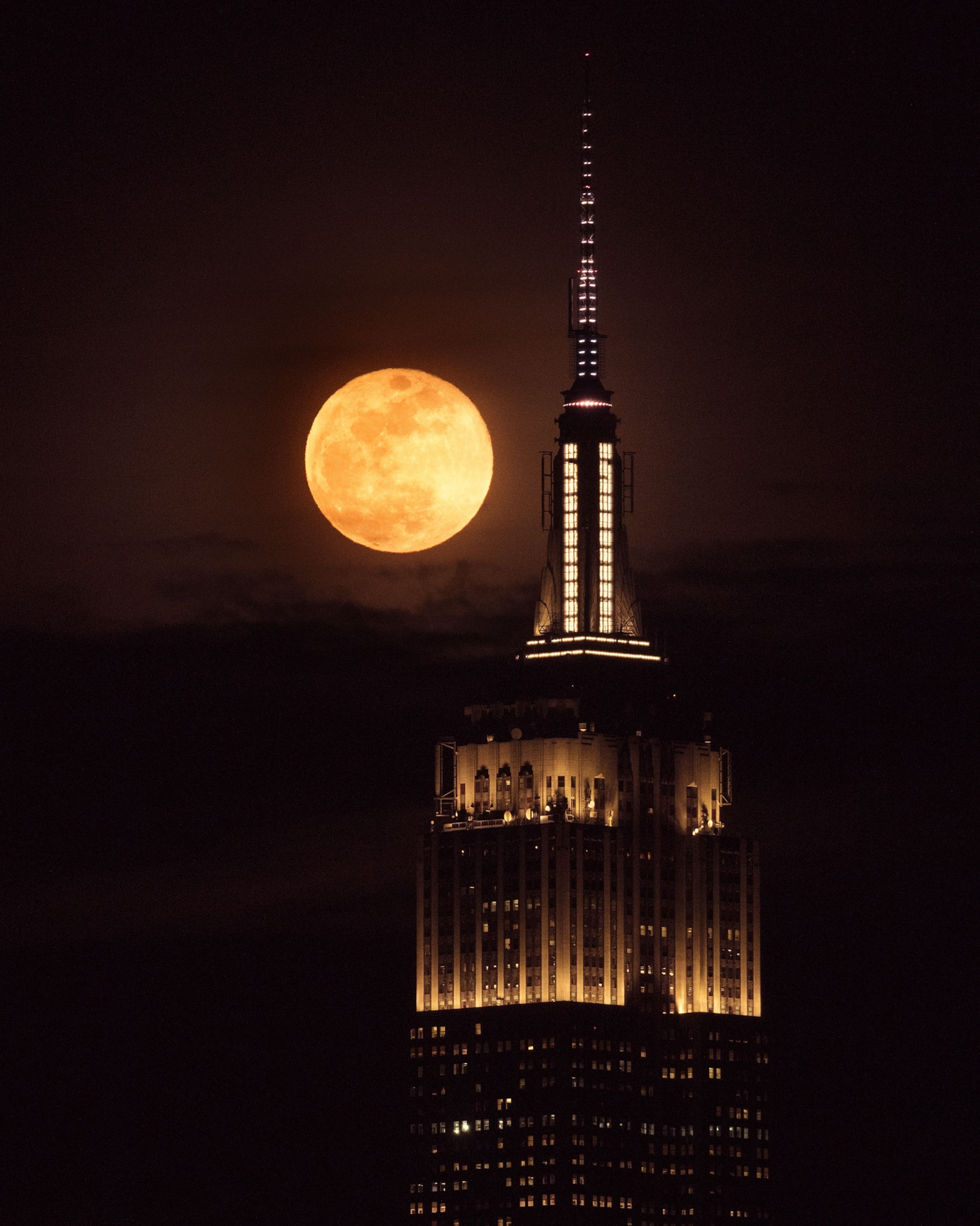 A full moon glows brightly beside the illuminated spire of the Empire State Building against a dark night sky.