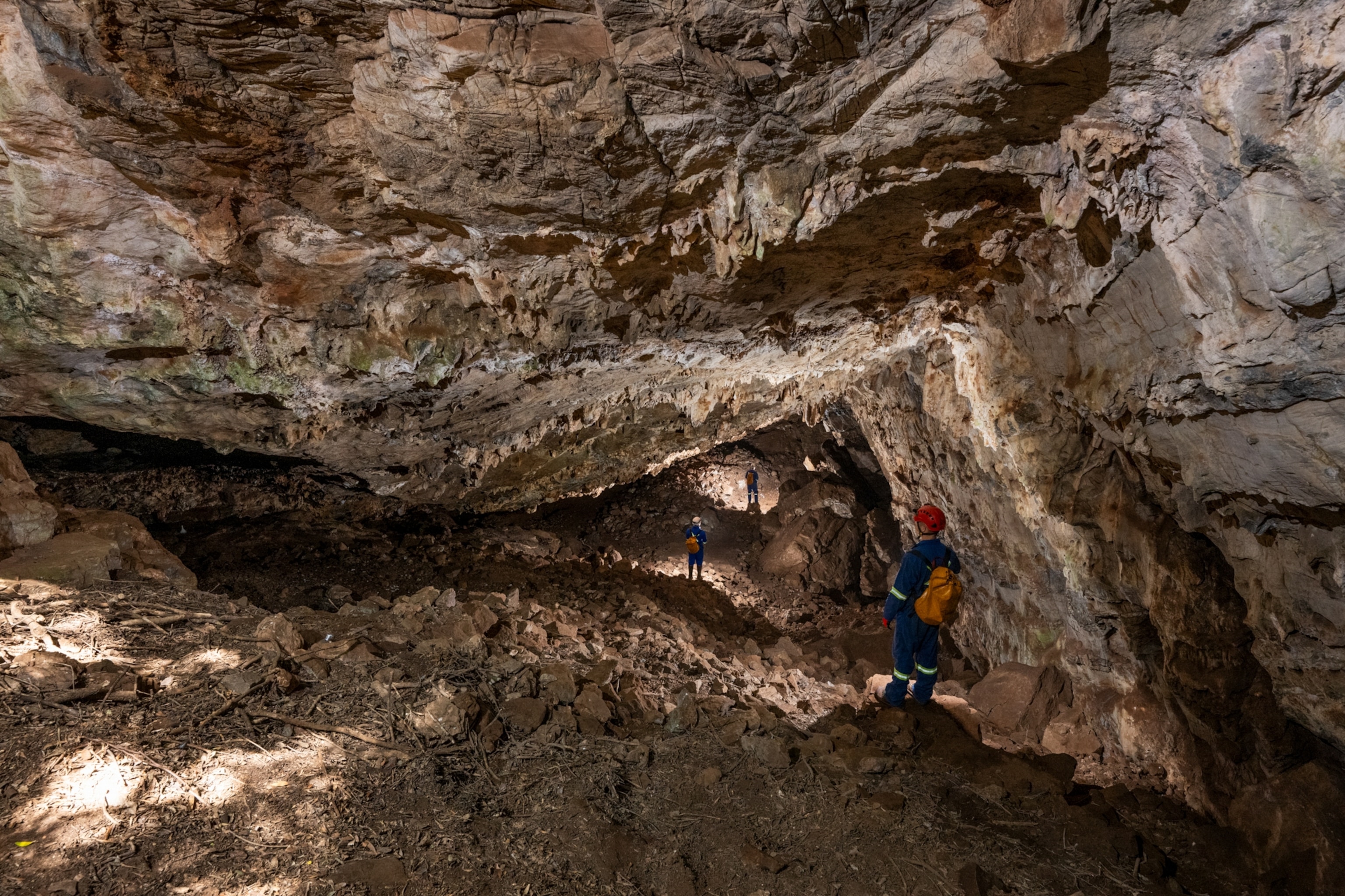 A rugged cave interior with three explorers wearing helmets and backpacks. The rough rocky ceiling is prominently visible.