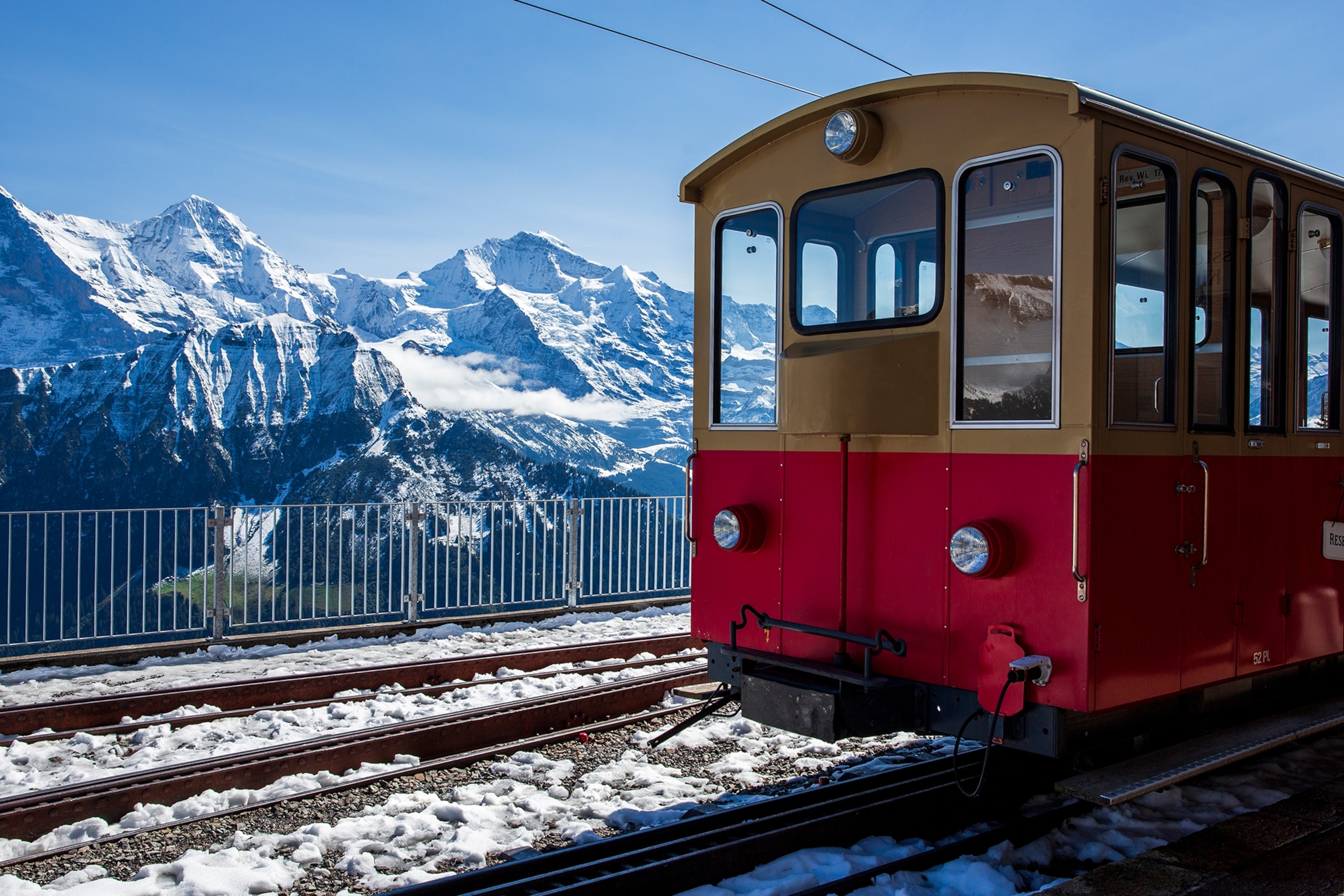 The front of a red train is pictured in the foreground, with snow-capped mountains in the background.