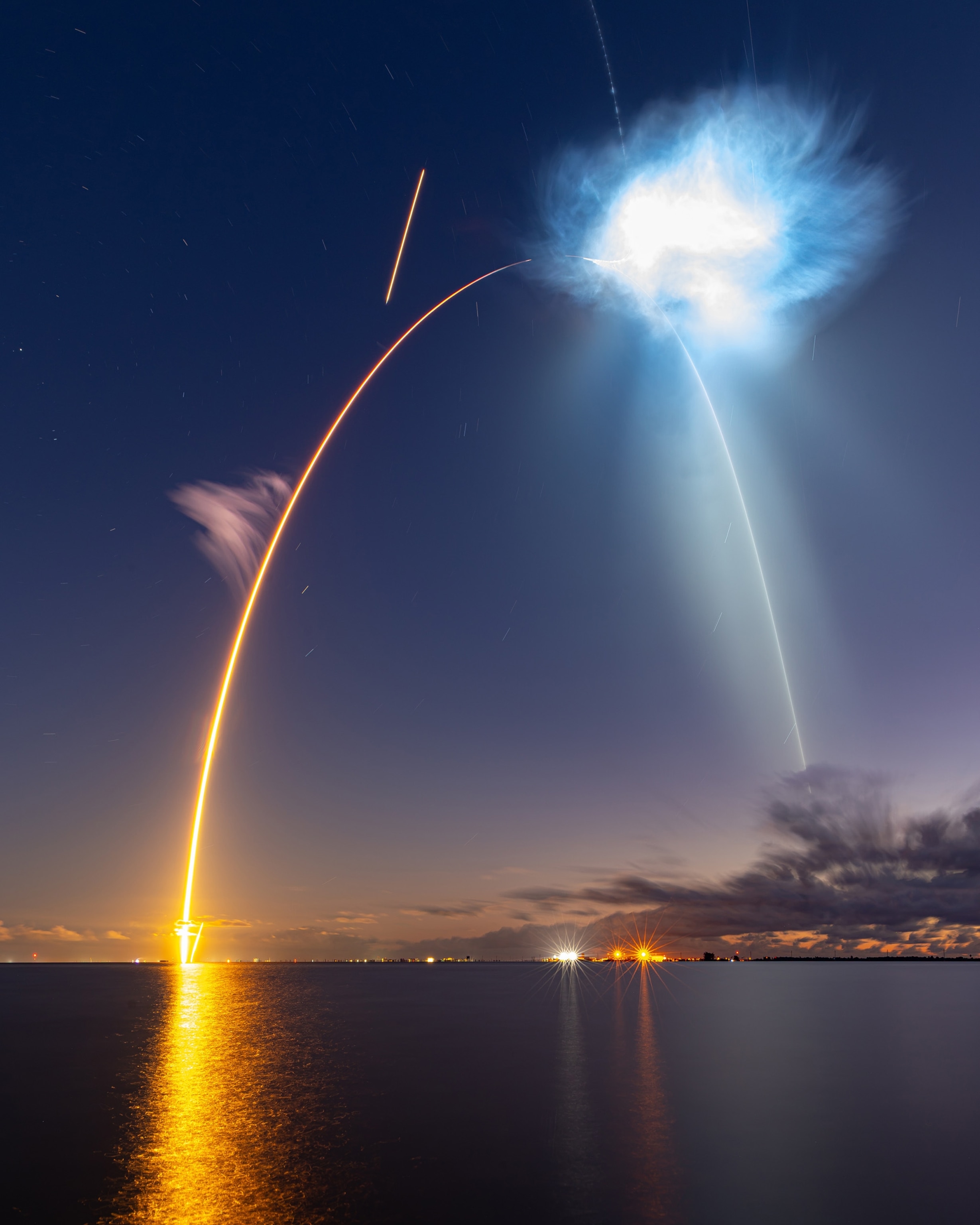 A SpaceX Falcon 9 rocket lifts off from a launch pad, creating a red and blue streak in the dark blue night sky above a dark body of water.