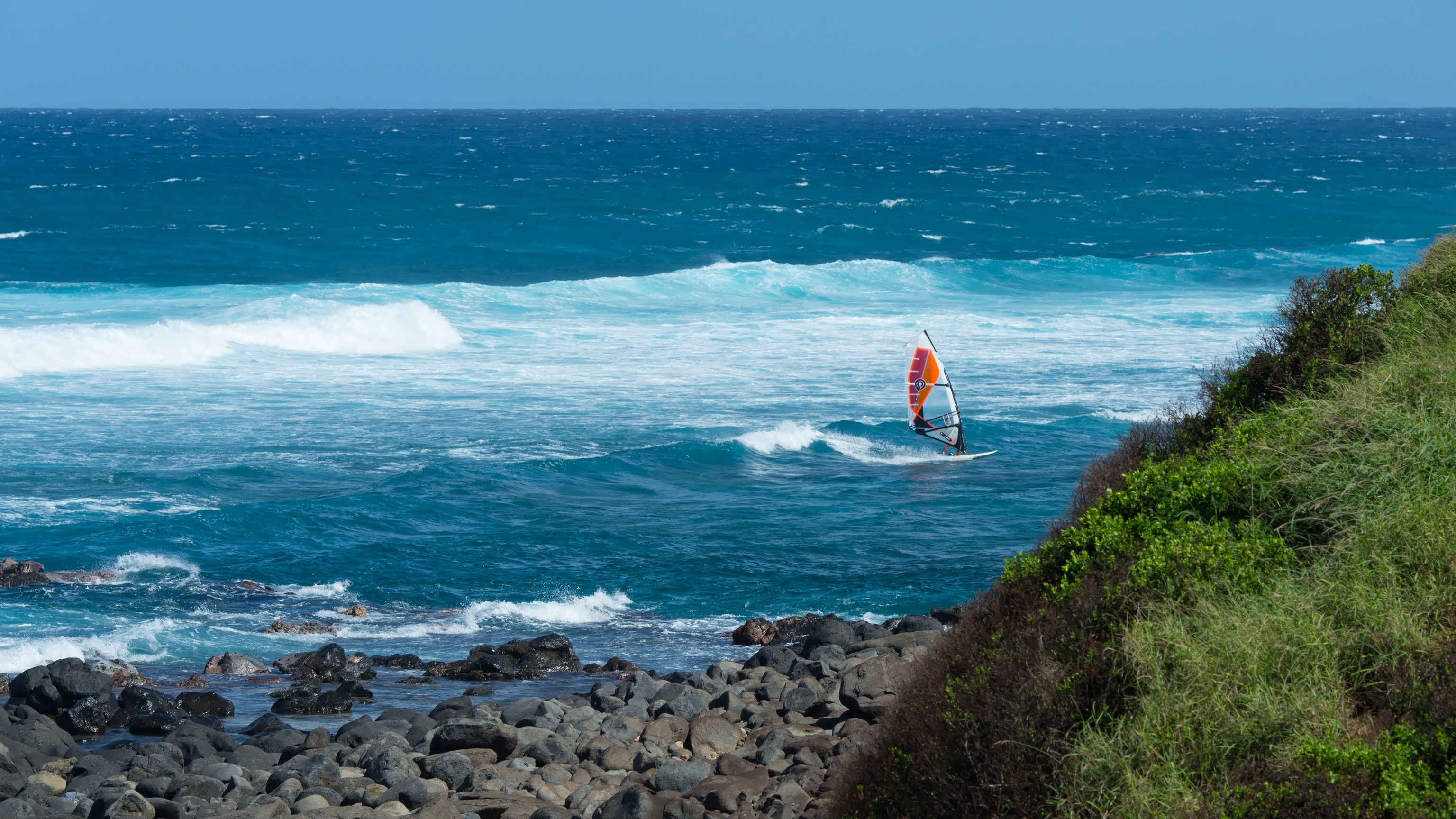 A kite surfer riding a wave in the ocean with a rocky and green covered cliff in the foreground.