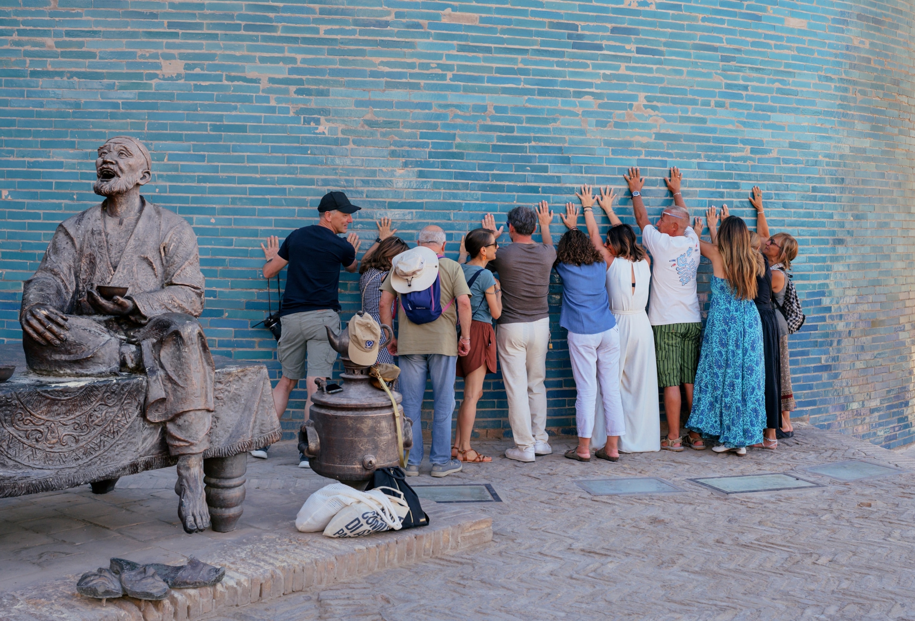 A group of 11 people stand facing a blue building wall with their arms stretched up and hands touching it. In the middle ground to left of the frame is a statue of a man with one leg crossed under his other leg which hangs straight down over a bench. In his hand he is holding a bowl and his mouth is open, as if talking or laughing, with his hand lifted slightly upward.