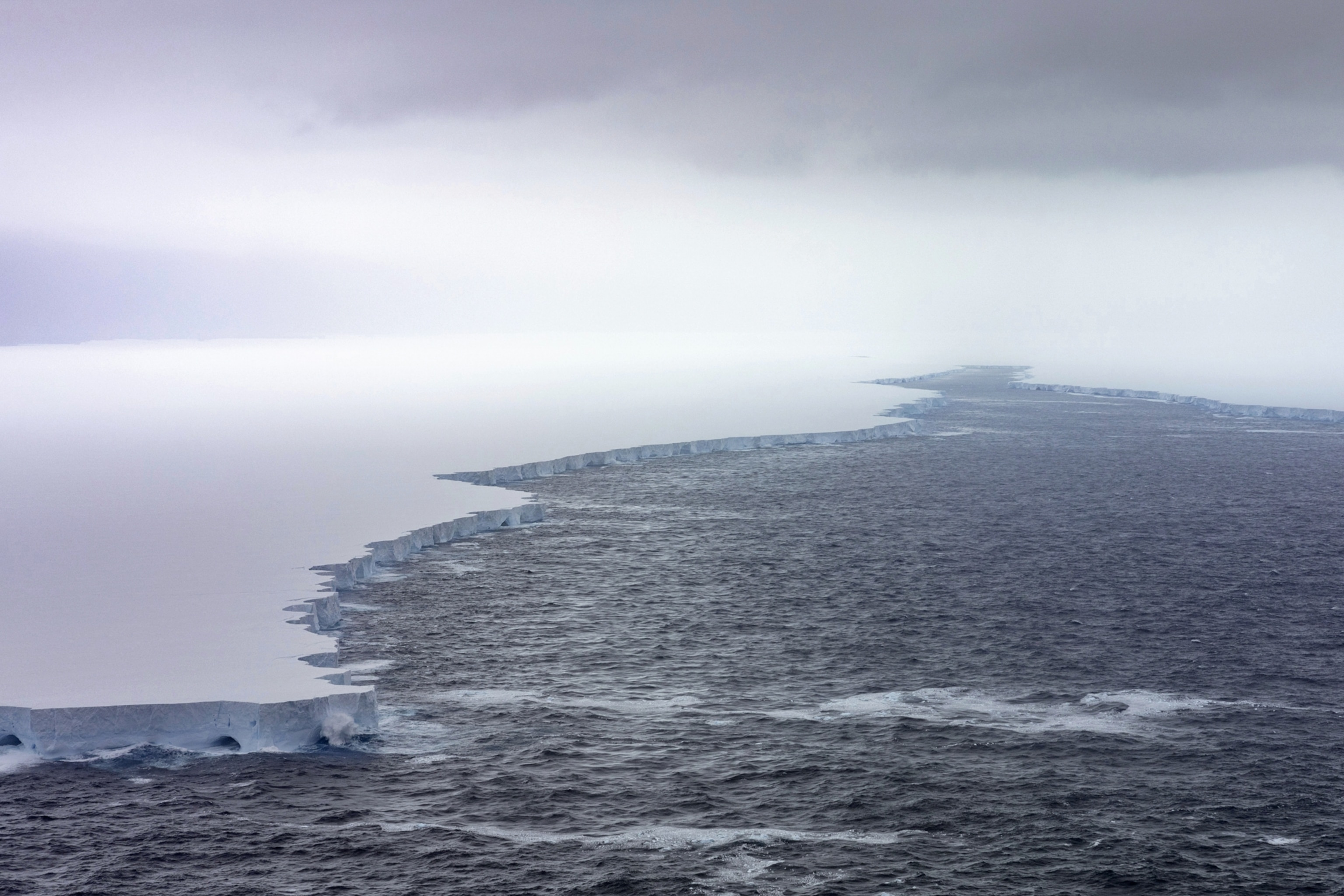 Expansive view of a massive tabular iceberg meeting the dark, choppy ocean under a cloudy sky.