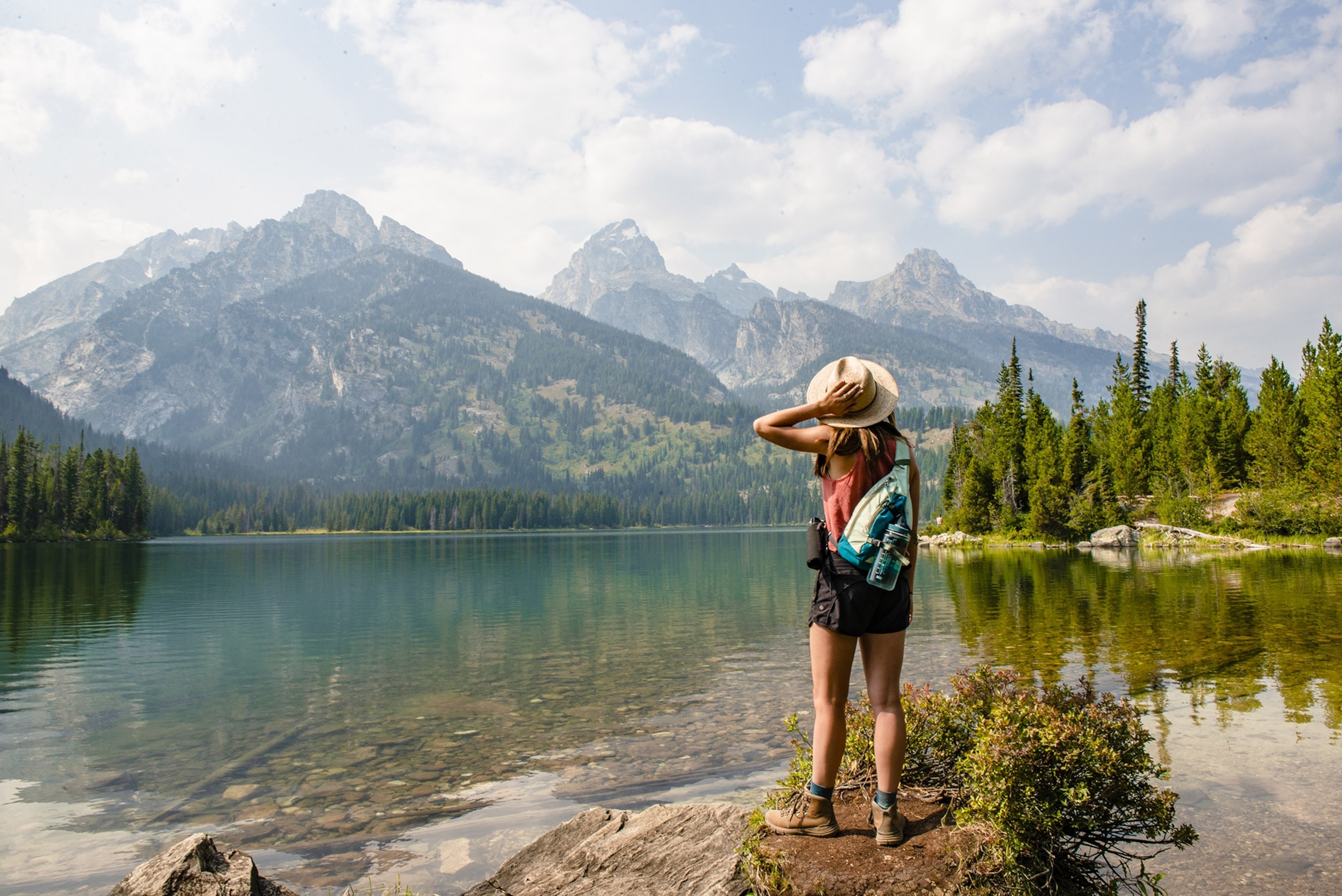 A woman faces away on the shore of a turquoise lake, looking up at a mountain range. She wears hiking boots and a hat.
