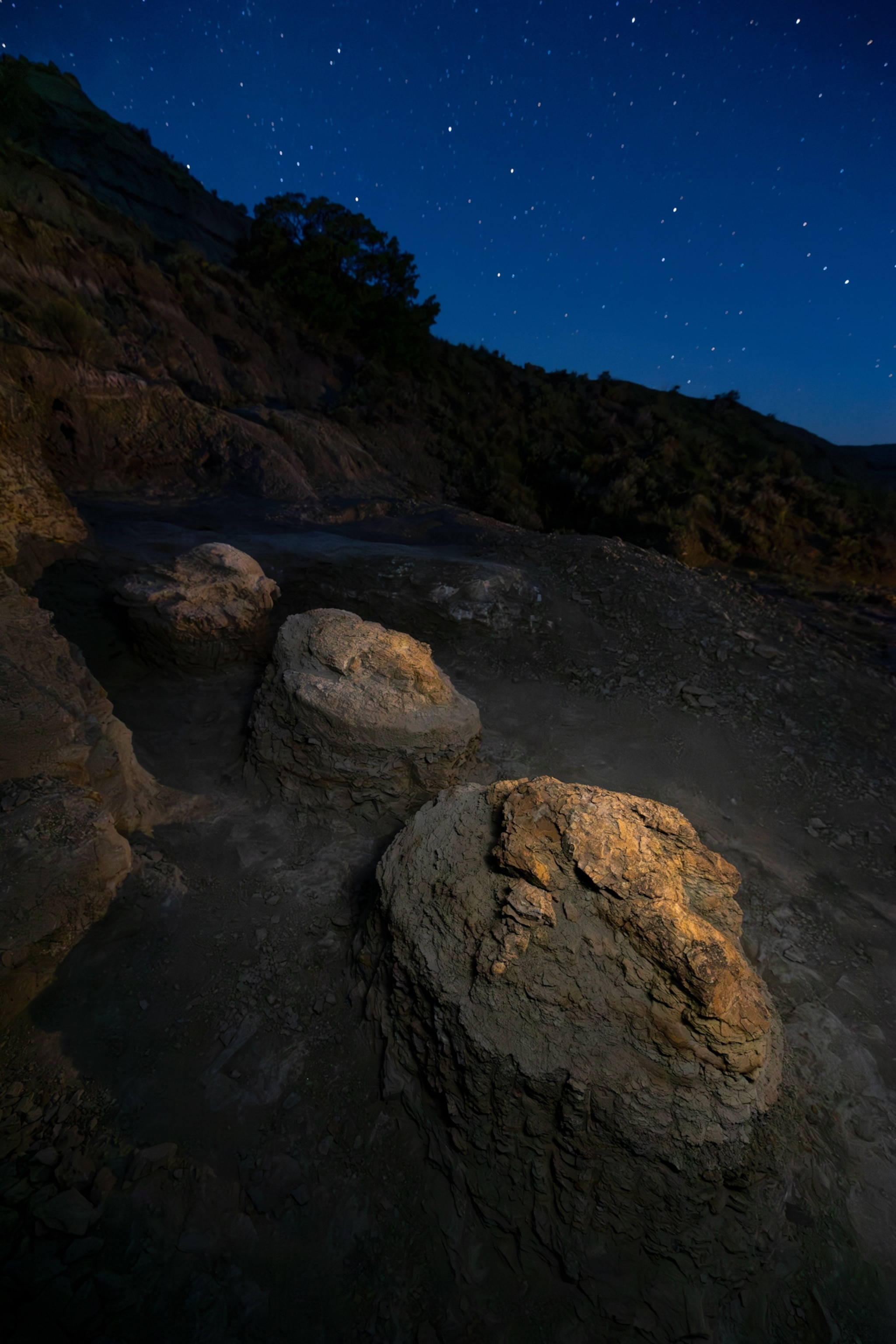 Star-filled night sky over a rugged volcanic landscape, with large fossilized footprints warmly lit in the foreground and a dark hillside beyond