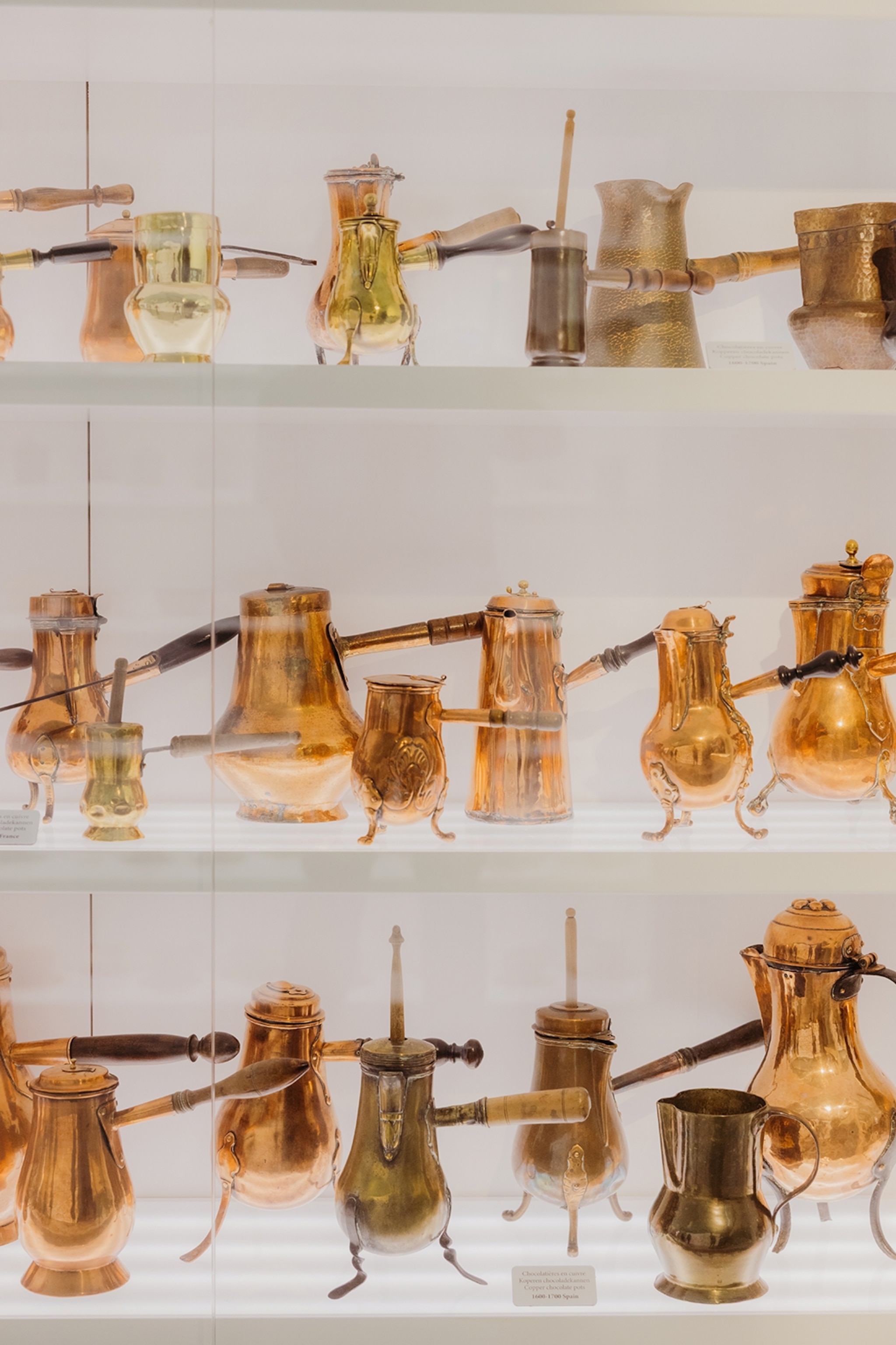 A close-up of a glass display in a museum with multiple shelves of brass chocolate pots.