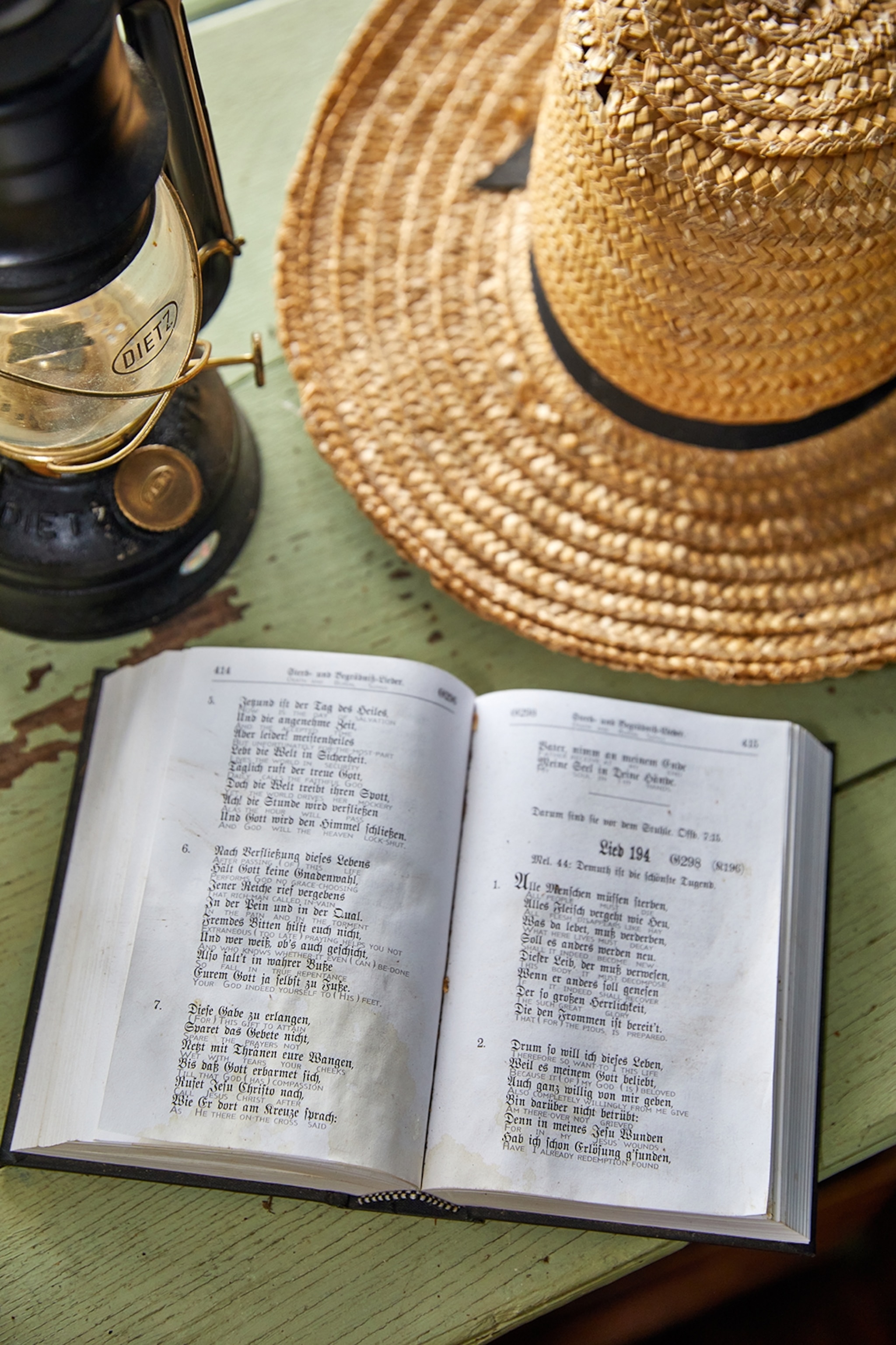 A close-up of an open hymn book on a rustic table with chipped paint, a straw hat and gas lamp.