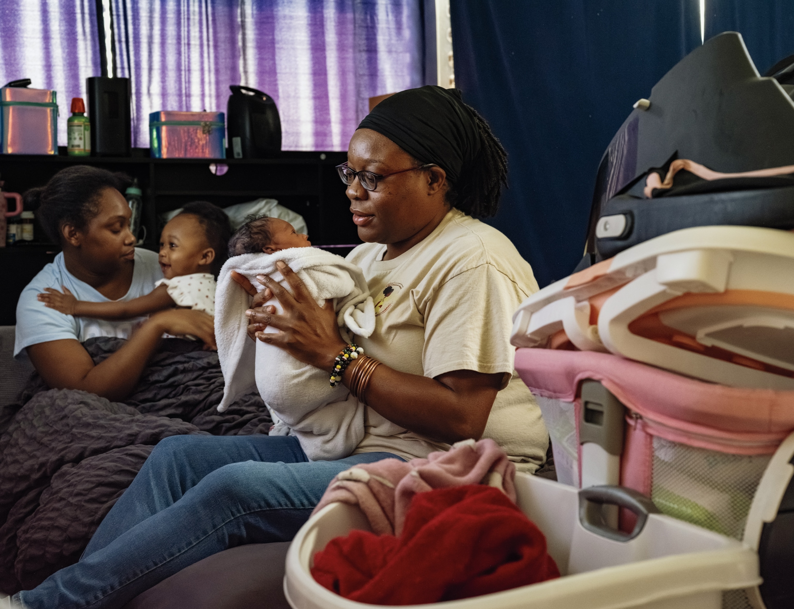 A woman sits and holds a swaddled baby, as another woman behind her holds a toddler. They are indoor and purple curtains cover the room's windows.