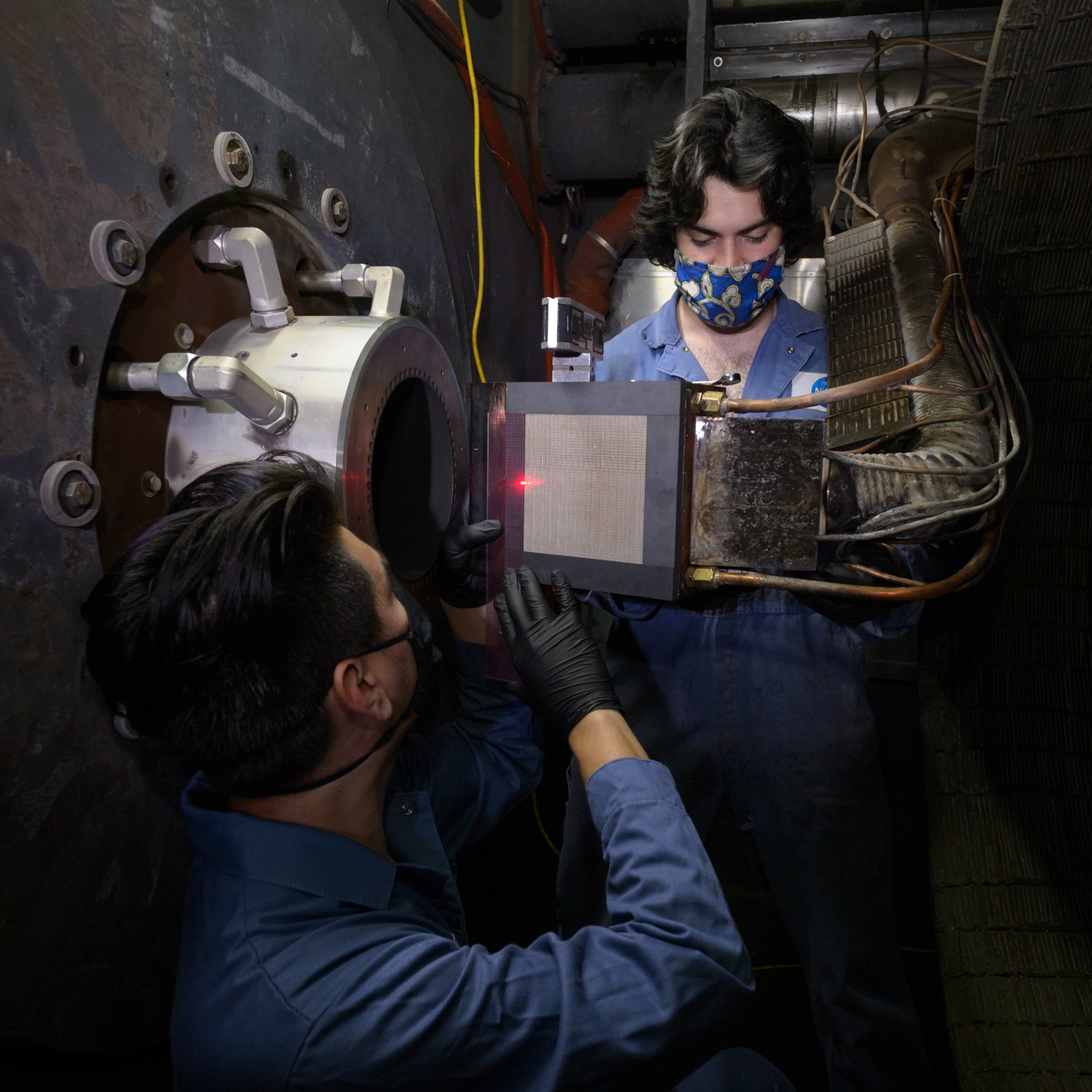 Two technicians in blue coveralls work inside an industrial setting, one holds a device with a red light while the other observes, both wearing face masks.
