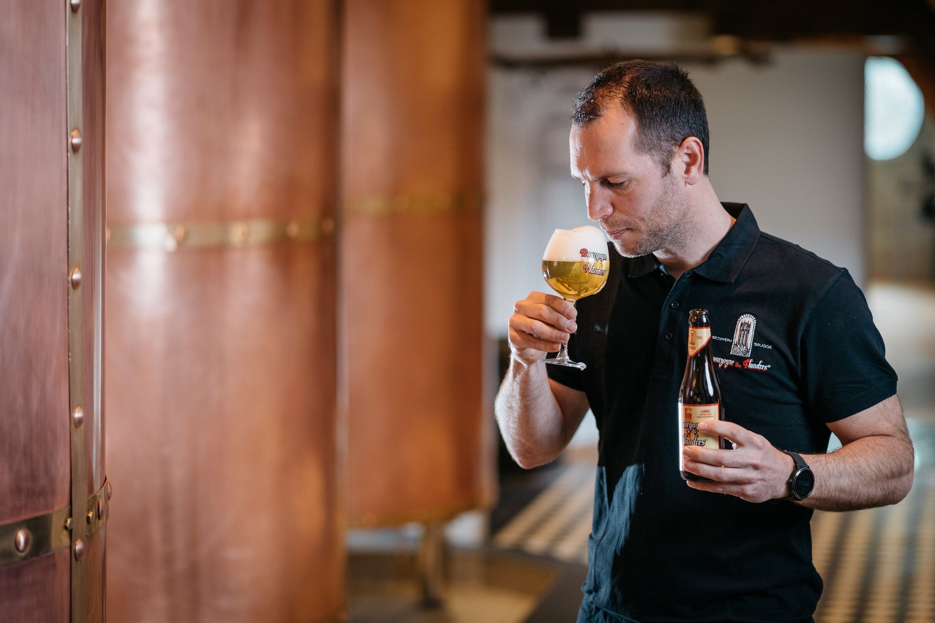 A man sipping a beer from a circular beer glass. It has a large head of foam, which almost touches his nose.