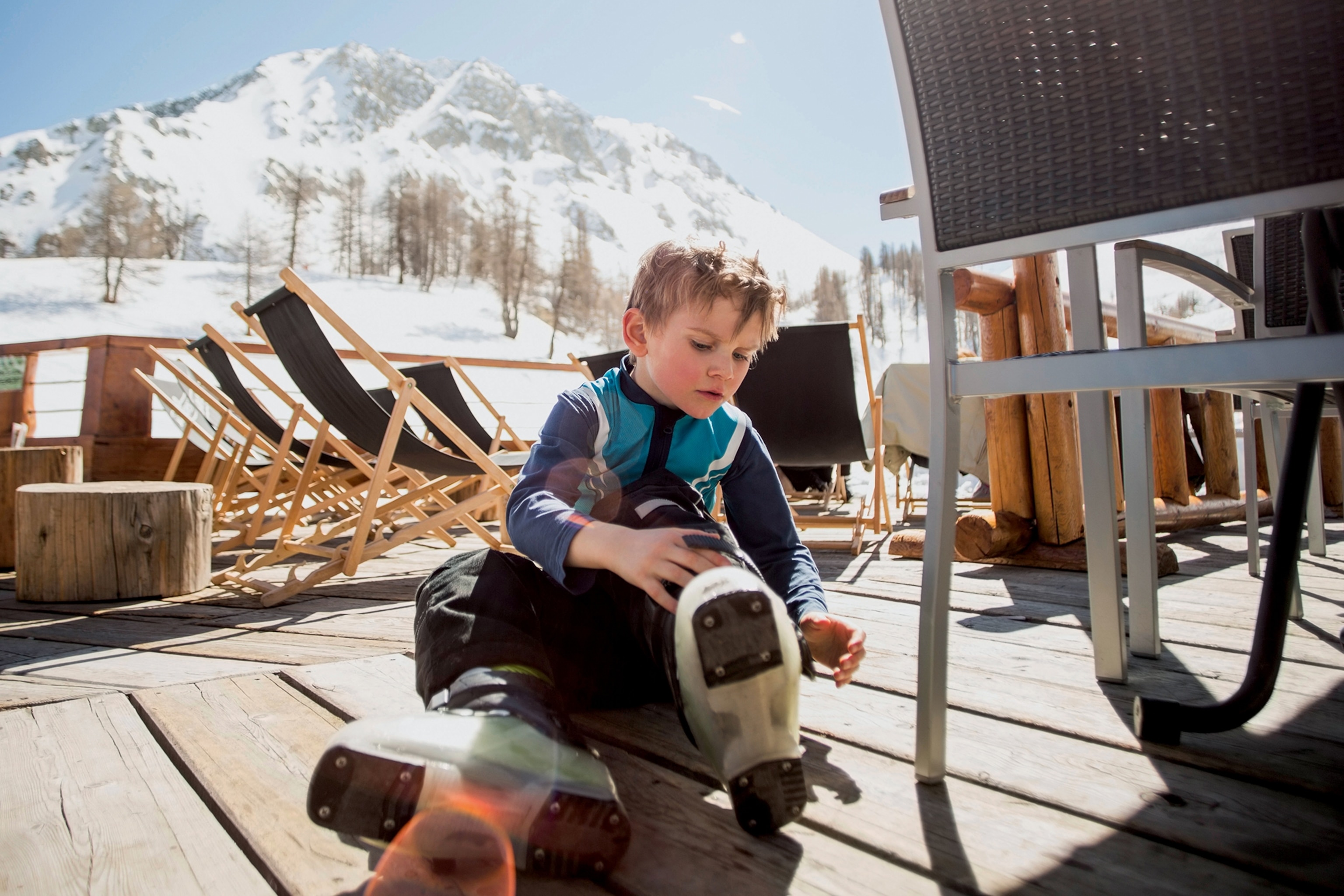 A small boy tying up his ski boots while sitting on a wooden terrace at the top of the mountains.