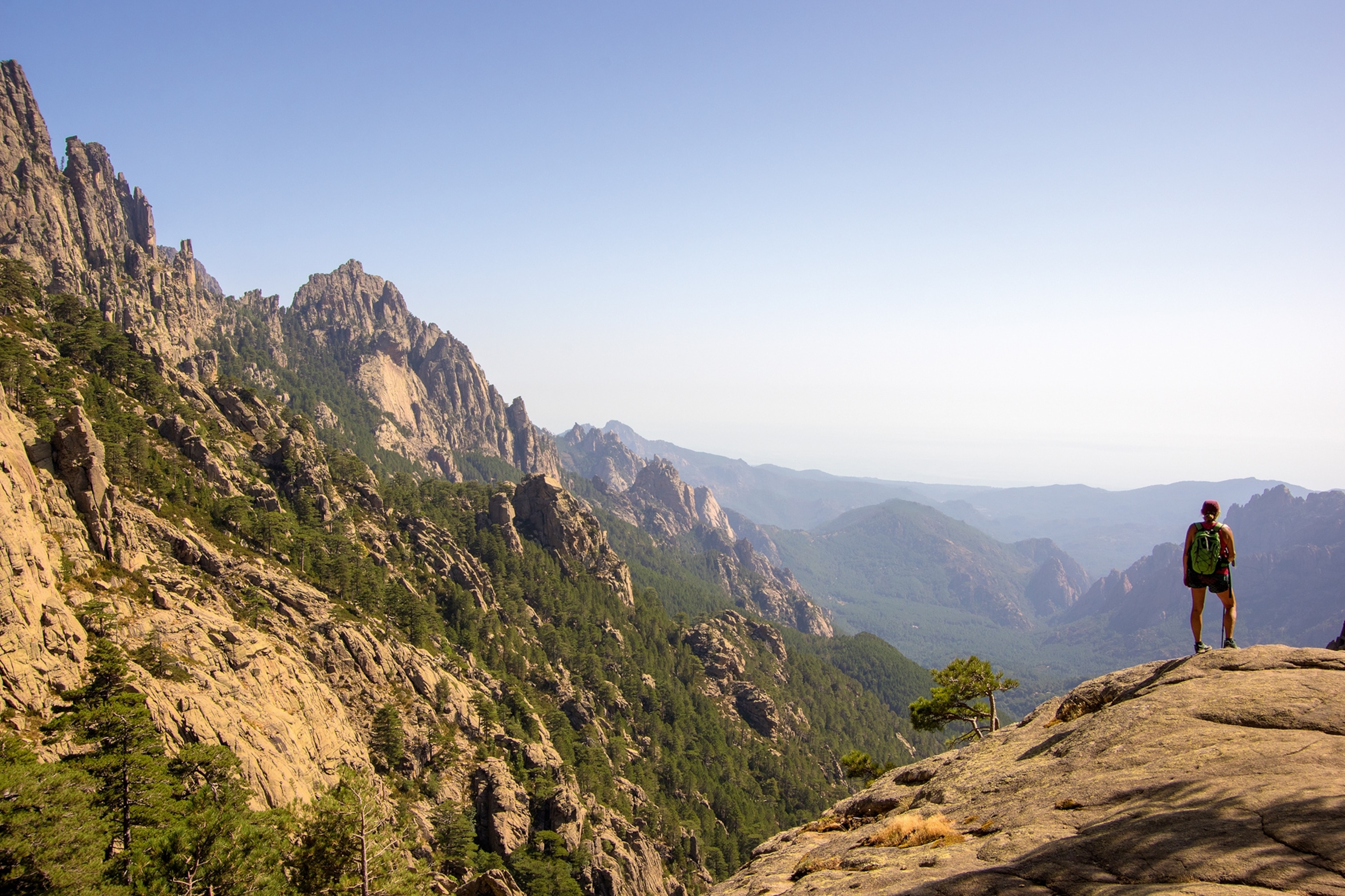 A hiker standing at the edge of a mountain cliff looking into a Mediterranean mountain valley.