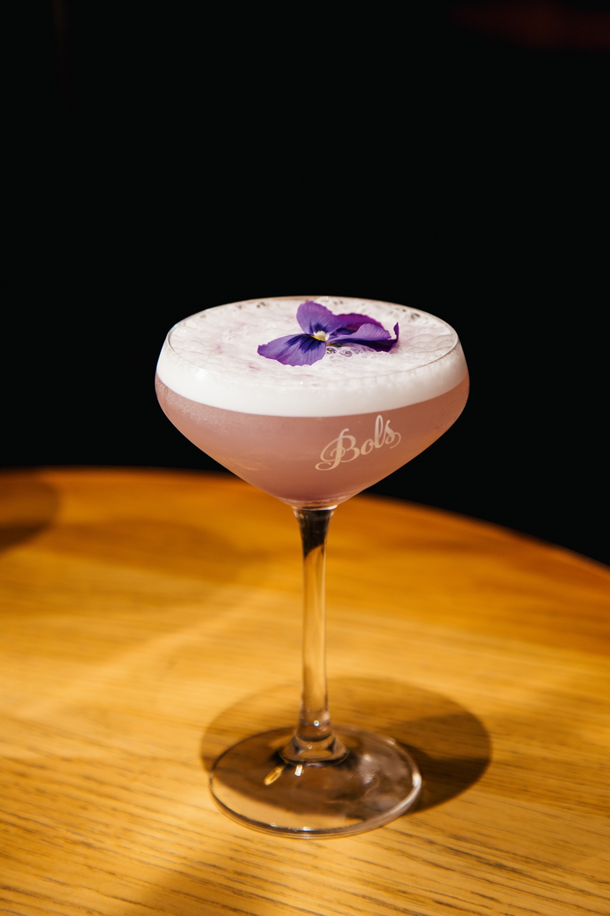 A close-up of a cocktail in a coupette glass with a foamy top and edible flower.