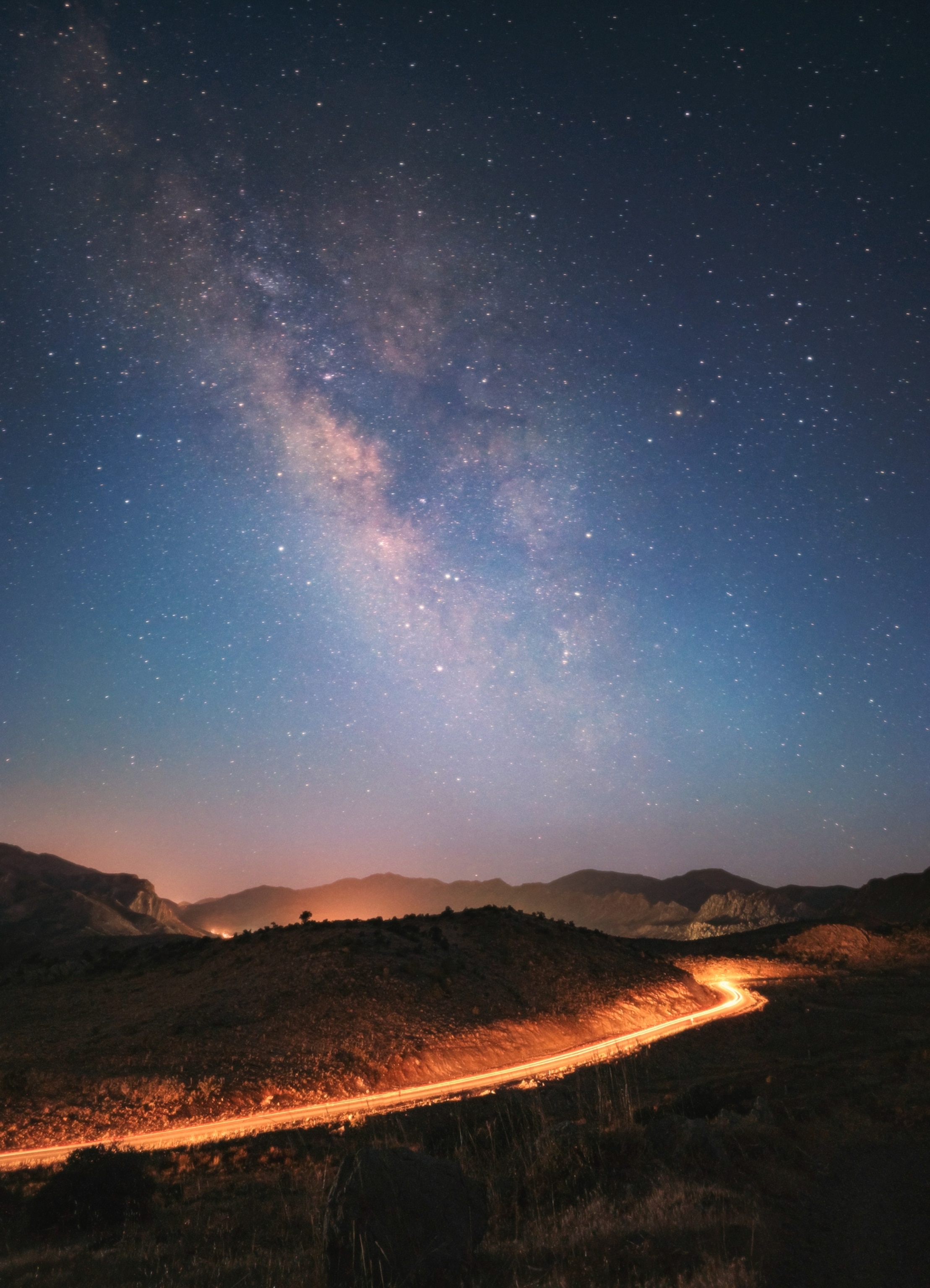 A starry night sky with a dense Milky Way galaxy band stretching over a winding, illuminated road through rolling hills