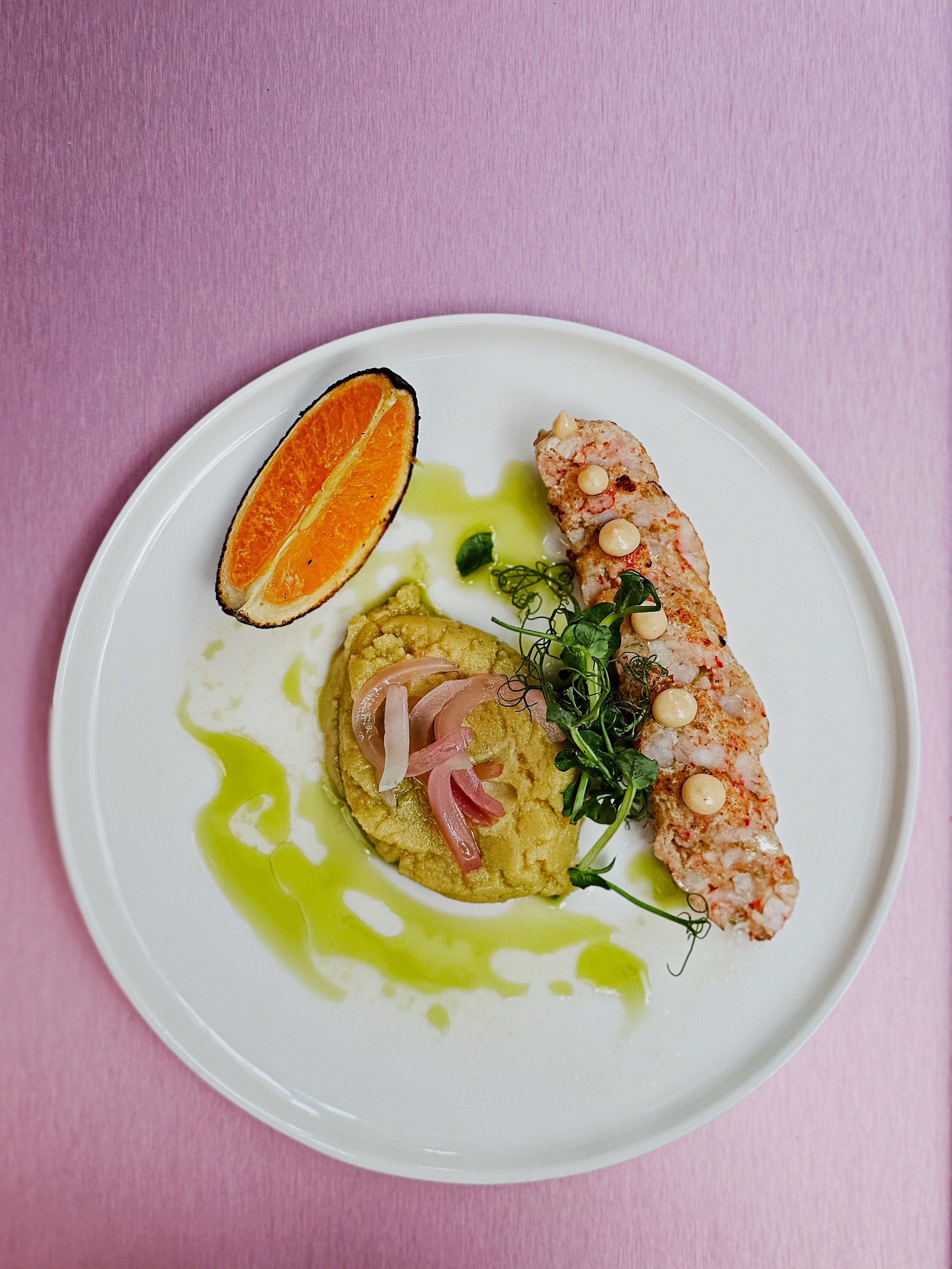 white plate with a pile of mashed fava, sausage, and various accoutrements on a pink background