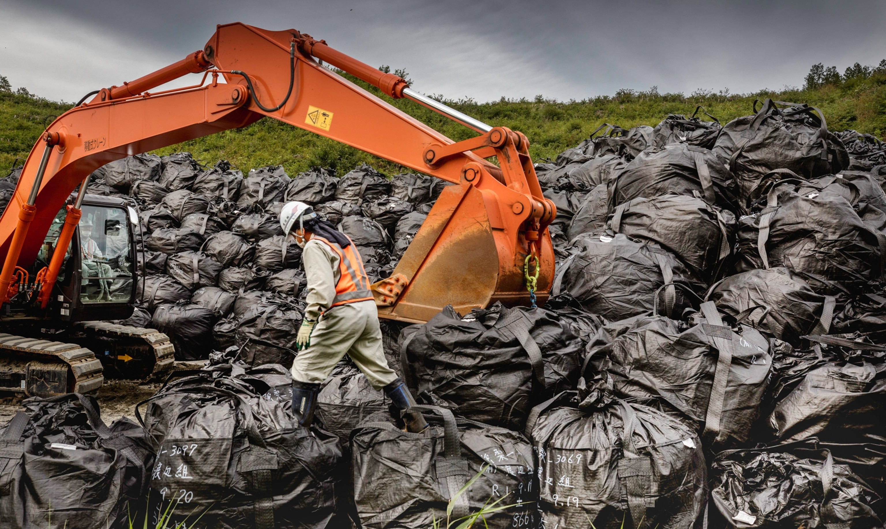 Person in rubber boots and respirator walking over piles of black bags.