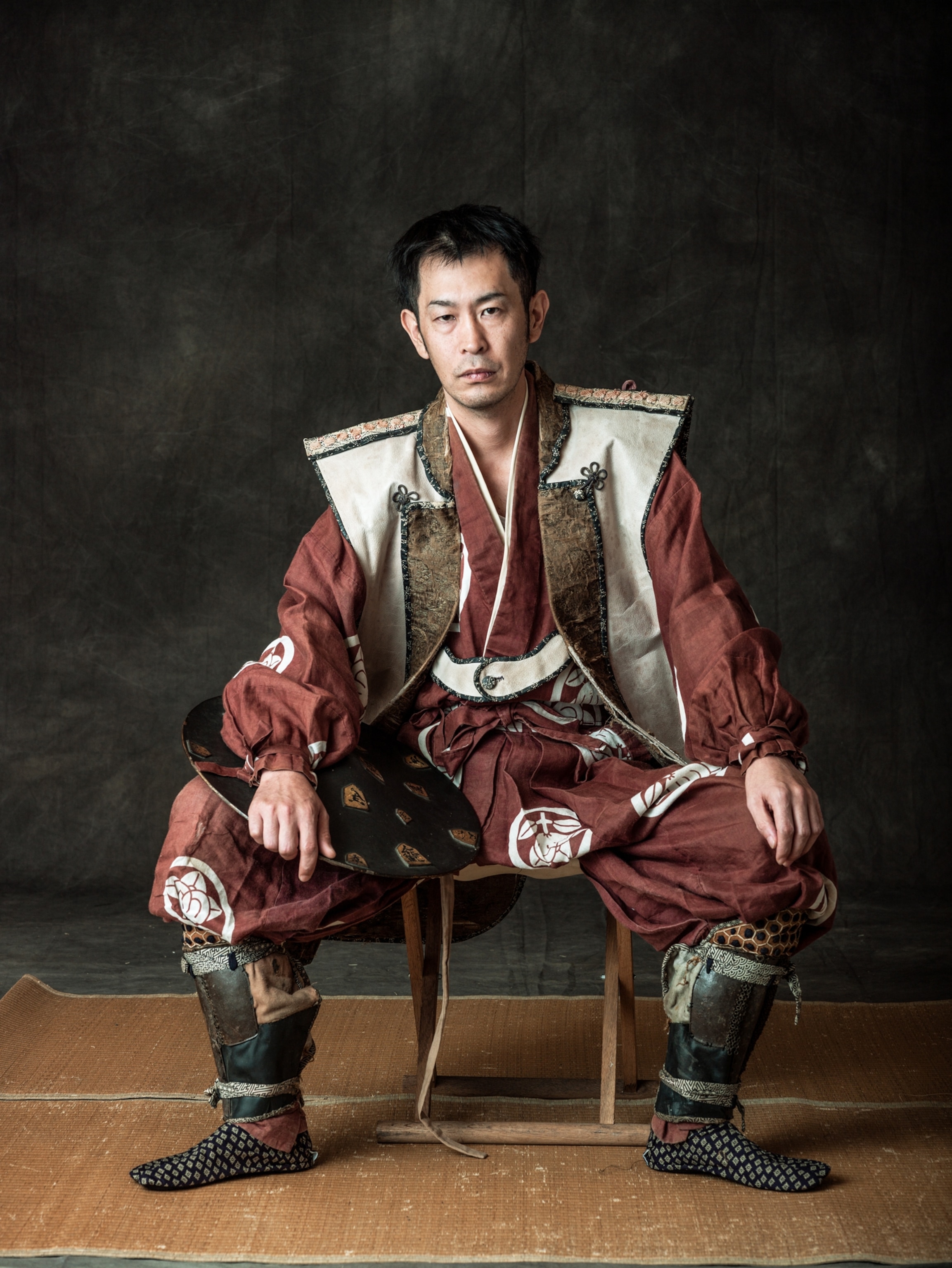 Picture of man in samurai costume without hat sitting on low stool with hand on his knees.