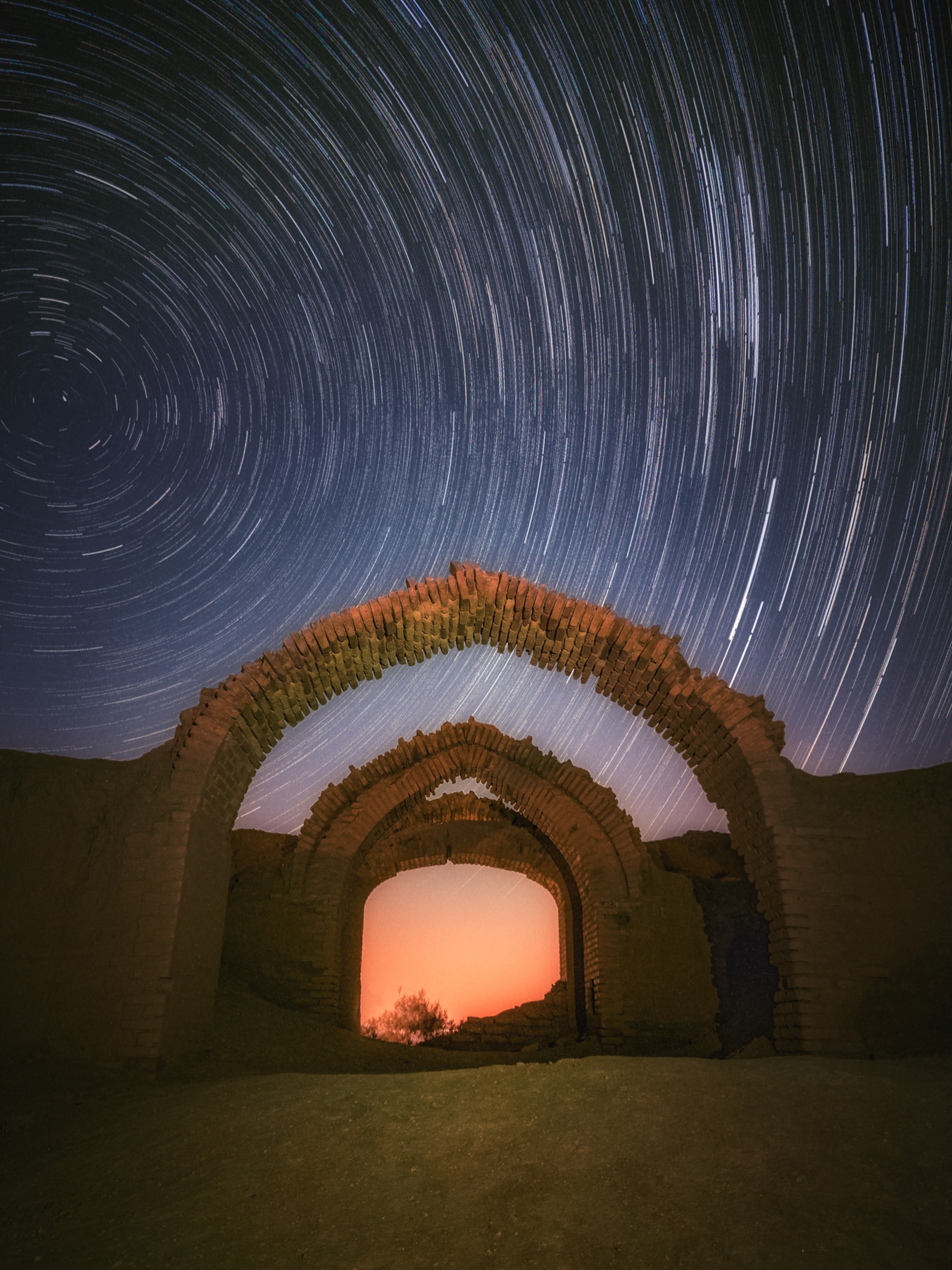 Ancient brick arches framing a soft orange sky on the horizon beneath swirling star trails above