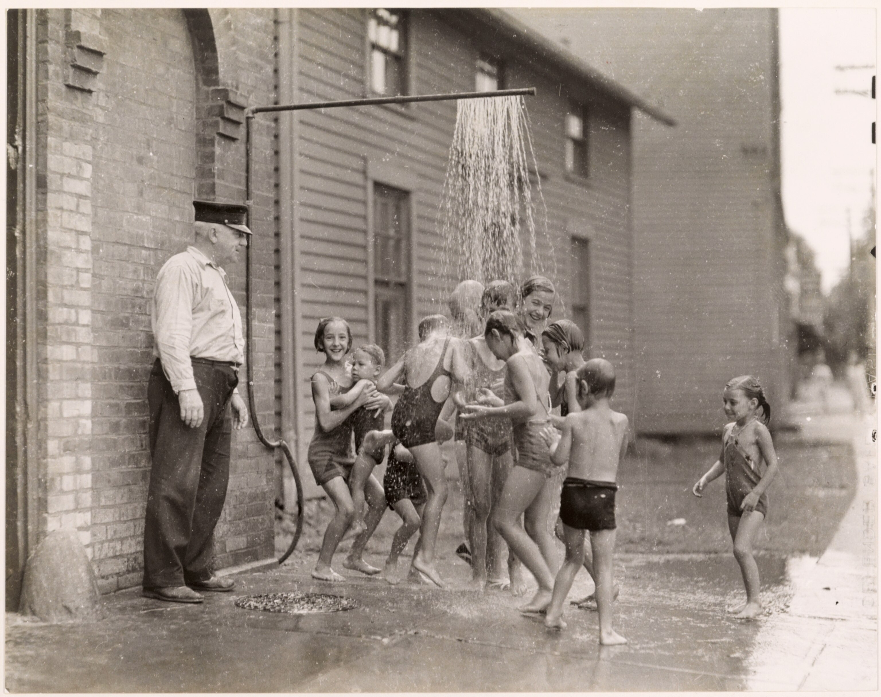 Children play under a pipe spouting water, 1935.