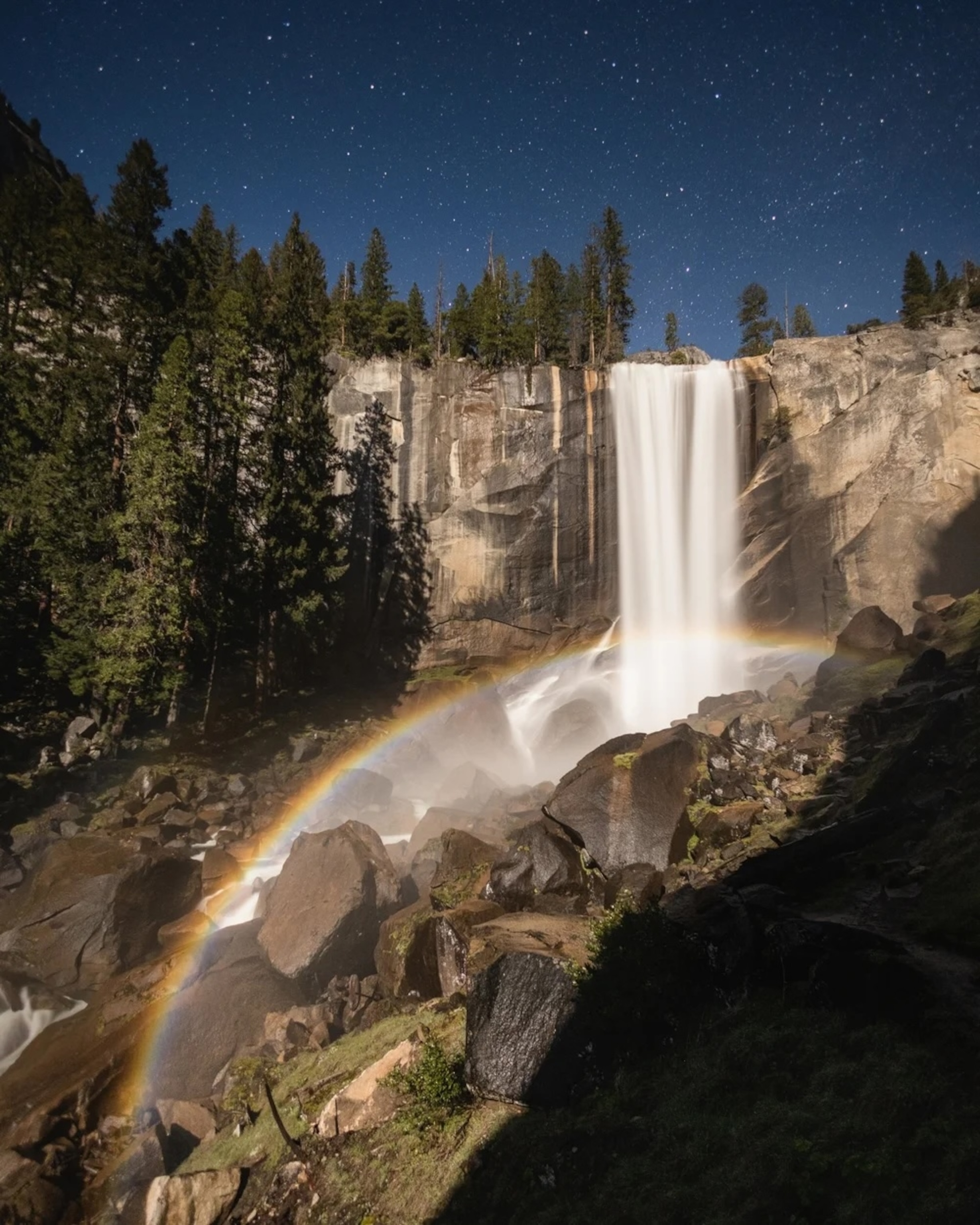 A tall waterfall flows down rocky cliffs at night, with trees above, stars in the sky, and a faint rainbow in the mist near the ground.