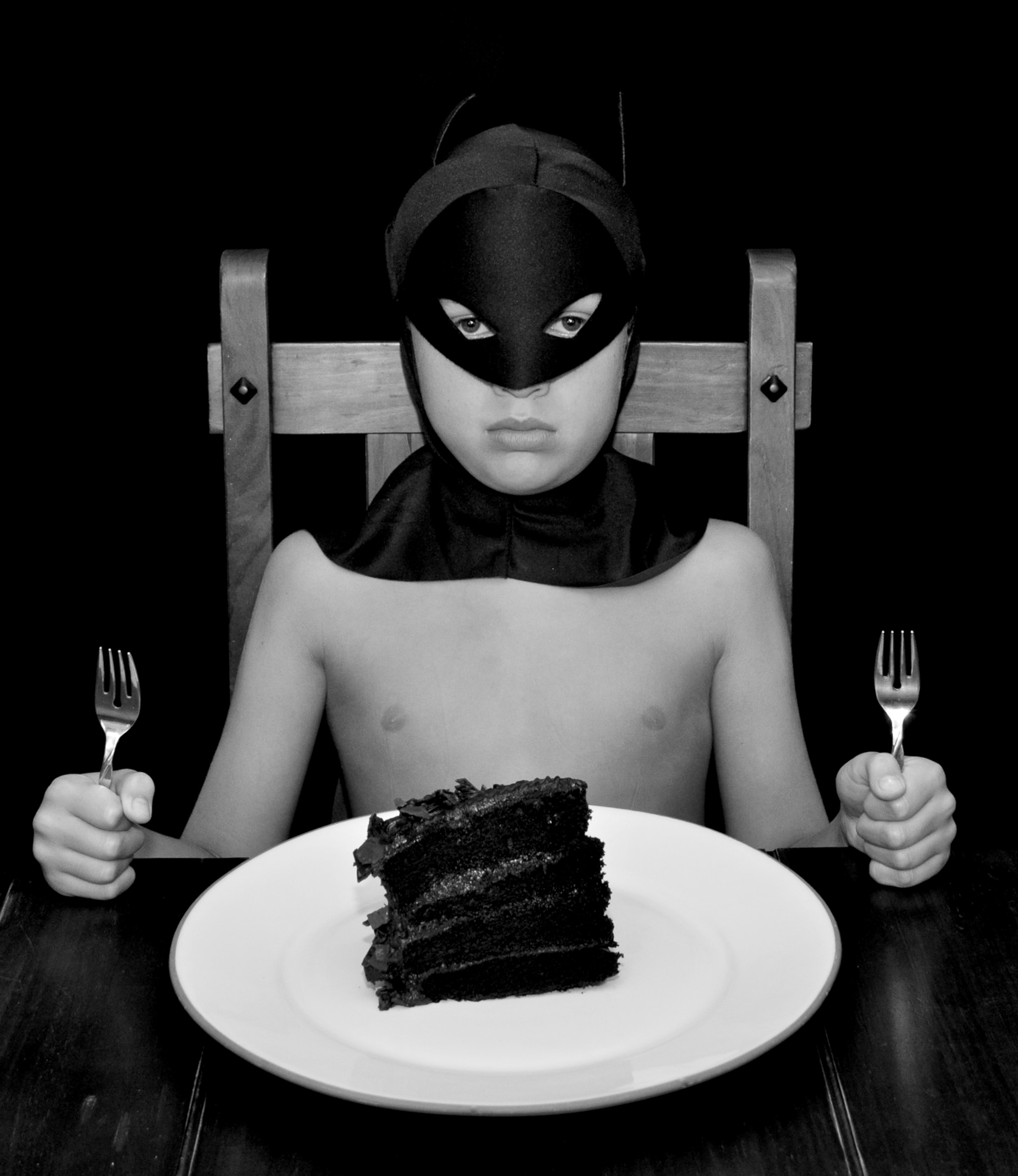 a young boy wearing a Batman costume and sitting at a table with a slice of cake.