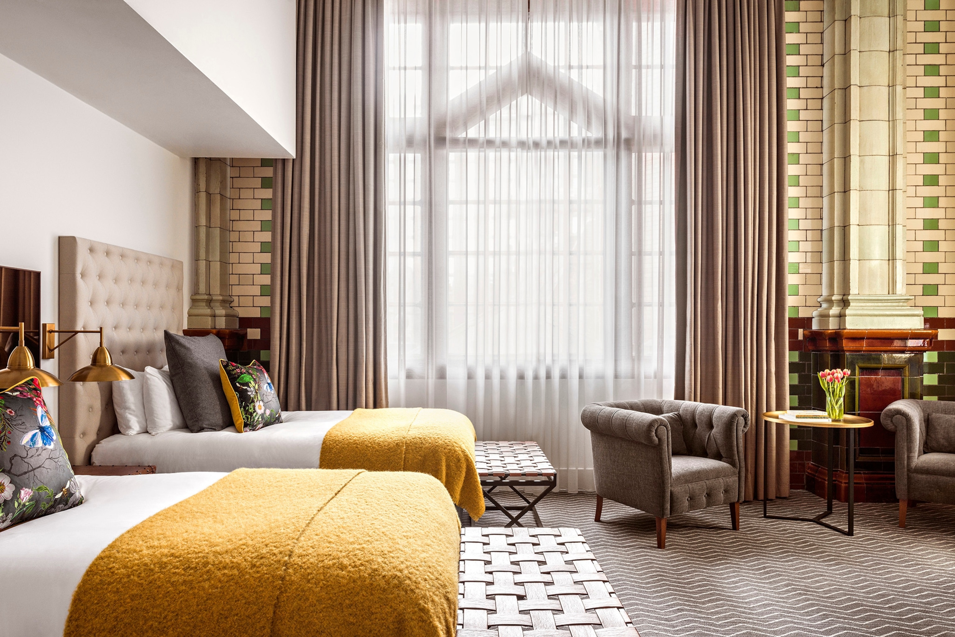 A hotel bedroom with double-height ceilings, brick wall features and two double beds in front of arched windows.