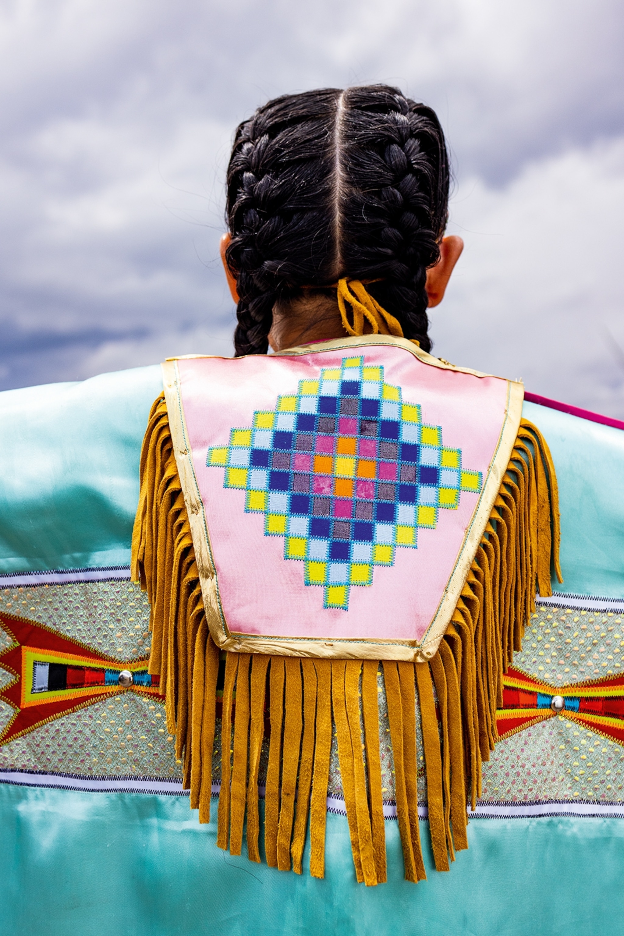 A girl with two braids shot from behind focusing on her beaded and tassled jacket.