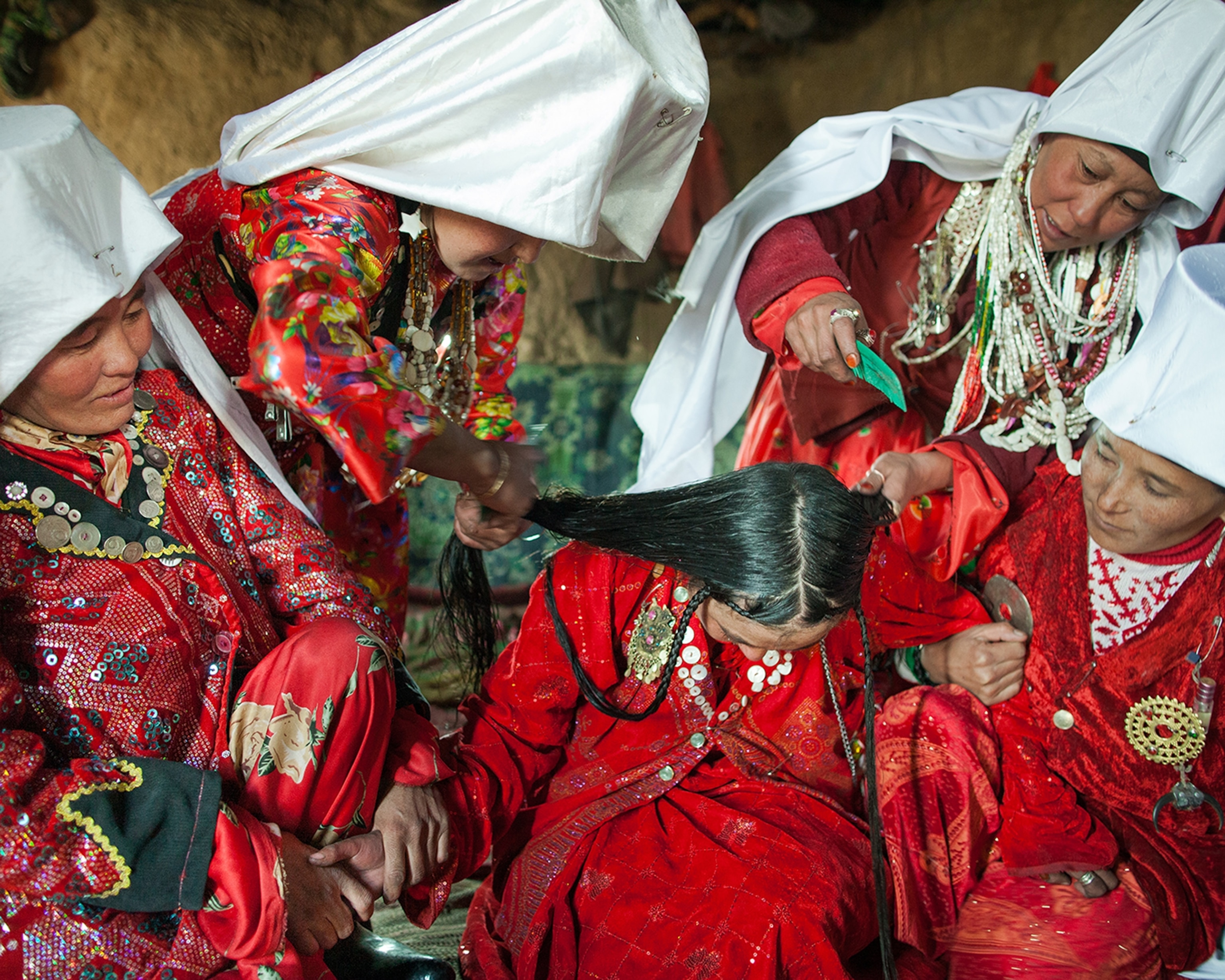 Four women surround a girl, who are all wearing red, and are combing her hair and holding her hand.