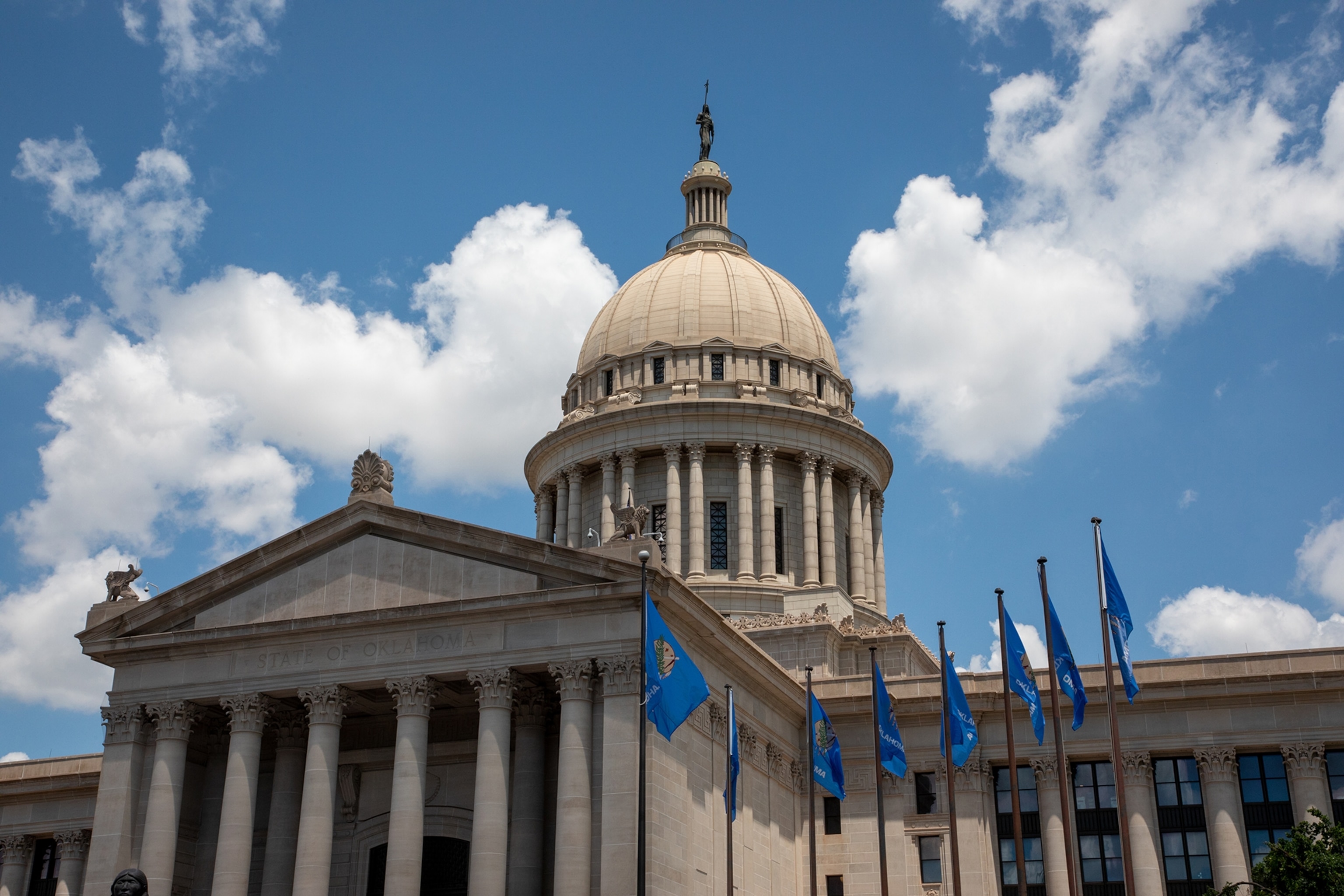 Blue Oklahoma state flags fly in front of the ornate Oklahoma State Capitol building.